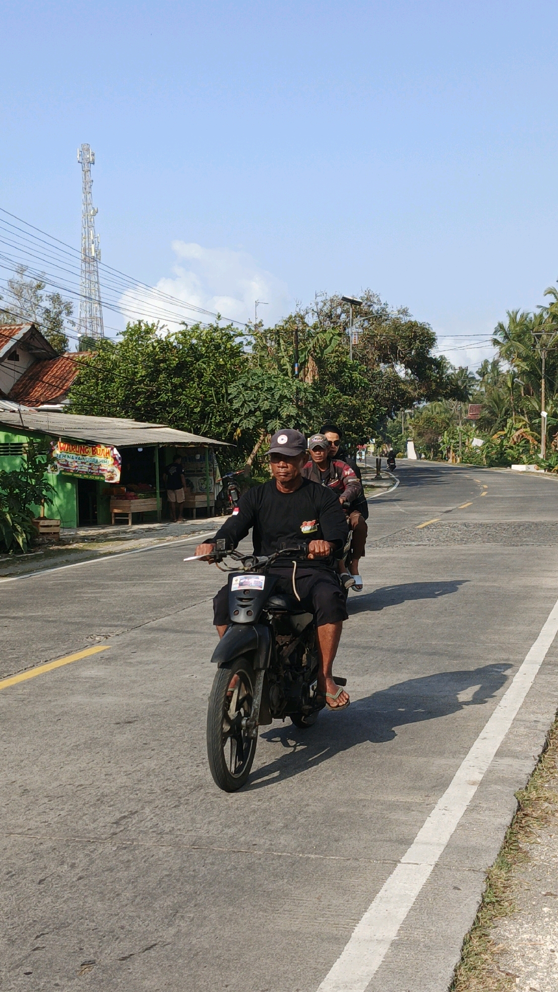 Three individuals are riding on a black motorcycle on a paved road. The person in the foreground, wearing a black long-sleeved shirt, black pants, and a baseball cap, is driving the motorcycle. They are looking forward. Behind the driver, a second person is seated, wearing a patterned jacket and sunglasses. The third person is seated behind the second, with their face mostly obscured.
The motorcycle is moving on the right side of the road, which has a yellow line on the left and a white line on the right. The road is surrounded by lush green trees and vegetation, with a few buildings visible on the left side of the road. One building appears to be a small shop with signage that reads "WARUNG BUAH" (fruit stall). In the background, a tall telecommunications tower is visible against a clear blue sky. The sun is casting shadows on the road, indicating it is daytime. The overall scene depicts a typical rural or semi-urban environment in Cibenda, Indonesia.