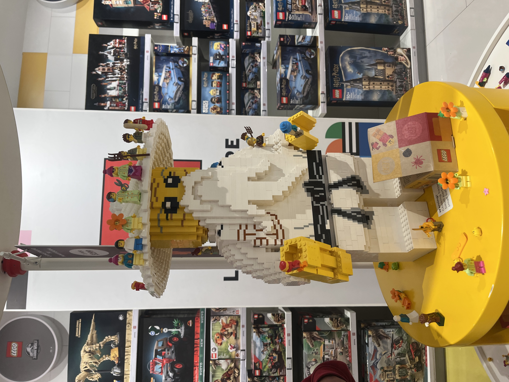 The image shows a large Lego sculpture of a person, likely a man, wearing a sombrero, surrounded by Lego sets on shelves in what appears to be a Lego store. The sculpture is made of white, yellow, and black Lego bricks, with miniature Lego figures placed around the sombrero and on a yellow platform connected to the sculpture. The shelves behind the sculpture display various Lego sets, including those from the Harry Potter and Jurassic Park series. In the foreground, the bottom of a face wearing a red beanie can be seen. The setting is identified as a Lego store, likely located in the city of Puebla, Mexico, based on clues from the packaging.
