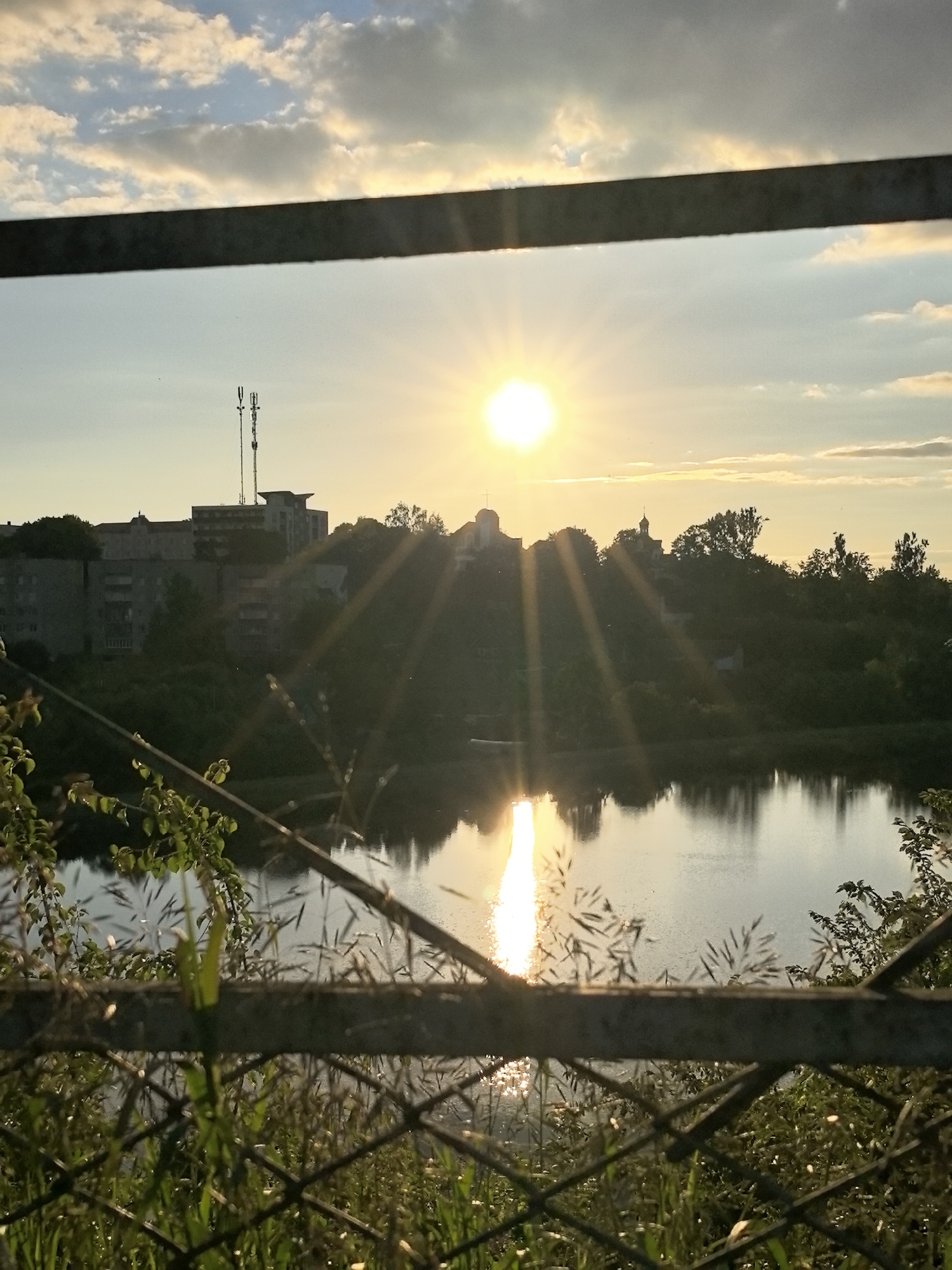 The image depicts a sunset over a body of water in an urban environment. In the foreground, a metal fence with vegetation in front of it partially obstructs the view. The fence is made of wire mesh and some horizontal bars. Beyond the fence, a calm body of water reflects the golden light of the setting sun, creating a bright streak of light on the surface. On the opposite bank of the water, a green, tree-covered hill rises, with buildings visible at the top. These buildings include what appears to be apartment blocks, a cell tower, and structures with domes, possibly churches or other religious buildings. The sky is a mix of blue and white clouds, with the sun shining brightly just above the horizon, casting rays of light downwards. The overall impression is a serene evening scene in a city.