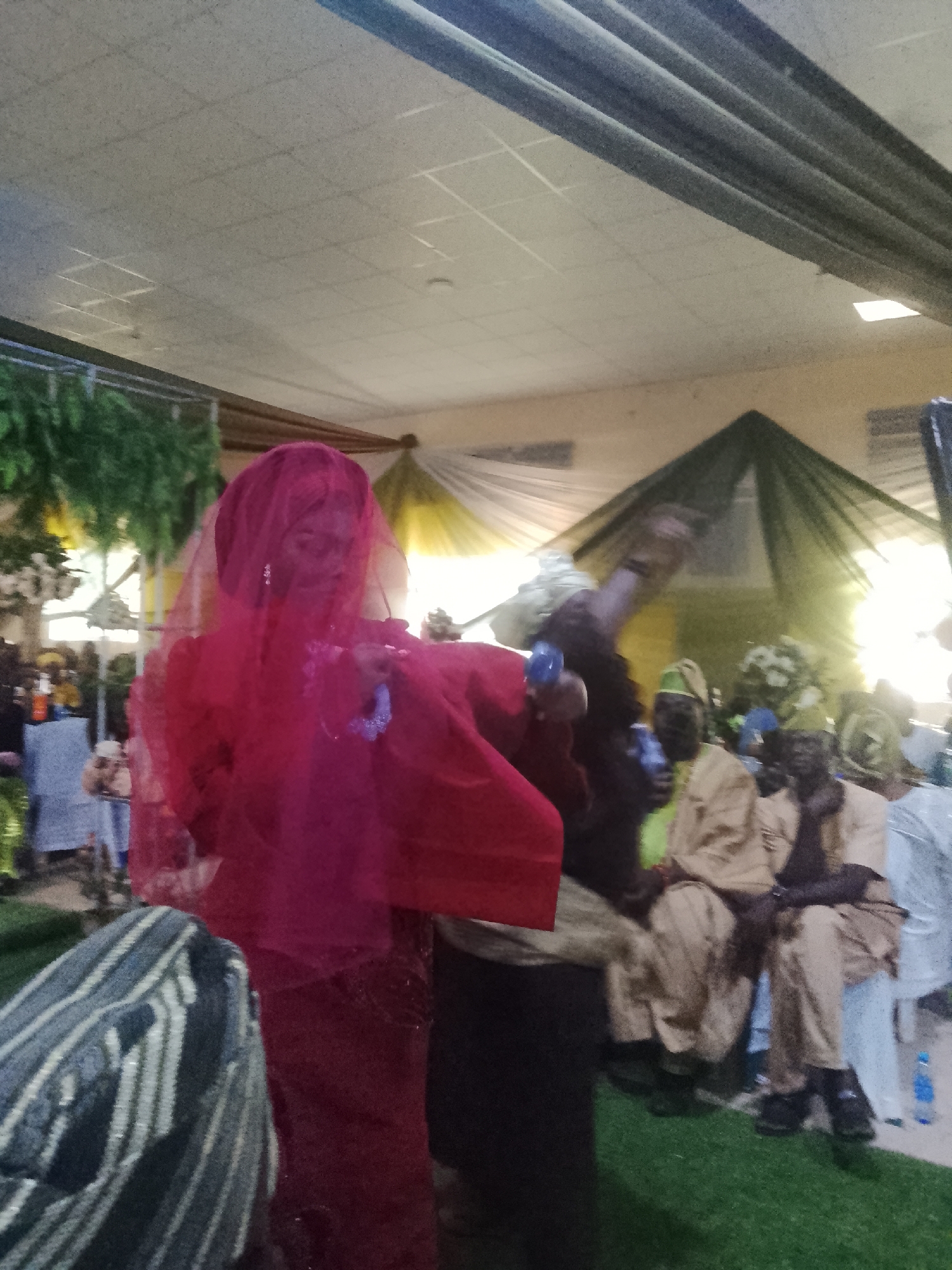 A woman in a red dress and veil stands prominently in the foreground of an indoor event space in Unknown, Unknown. Her features are obscured by the veil. To her right, a group of seated men, some wearing traditional headwear and tan clothing, observe the scene. Behind them is a backdrop of draped fabric in shades of yellow and green. Artificial turf covers the floor near the seated men. A water bottle sits on the floor to the right of the men. Other unidentified people are visible in the blurred background to the left, along with floral decorations. The ceiling is visible above, with visible vents and overhead lighting.