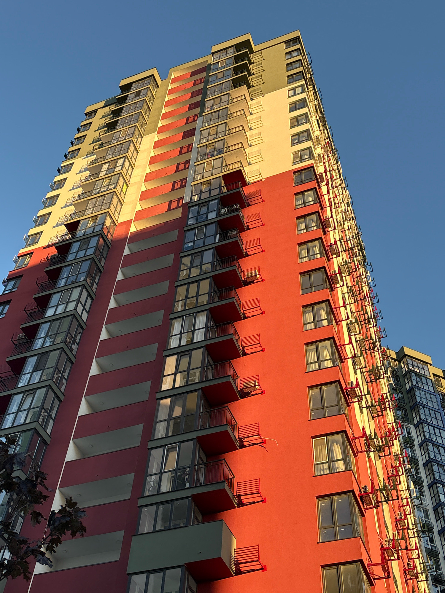 A low-angle shot captures a modern, tall residential building against a clear blue sky in Kyiv, Ukraine. The building features a striking facade with alternating bands of red, cream, and a pale green-yellow, illuminated by the warm, golden light of the setting sun, creating strong shadows. Many balconies with dark railings are visible, some with small chairs. Numerous air conditioning units are mounted on the exterior walls. The building is seen from a low perspective, emphasizing its height and the deep blue of the sky.