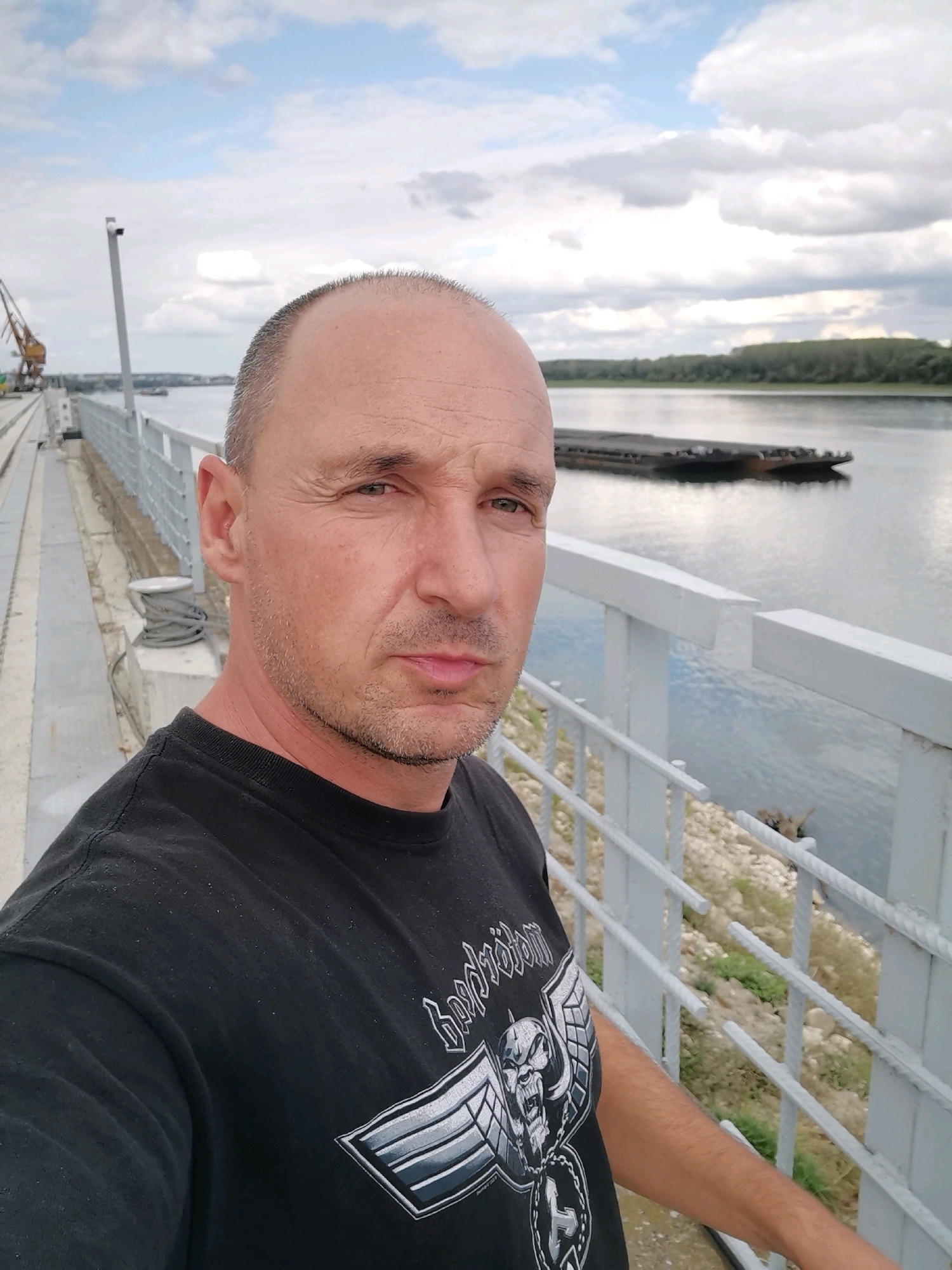 In a medium shot selfie, a bald, middle-aged man with stubble stands on a pedestrian walkway beside a river. He is wearing a black t-shirt with a graphic design of a skull and wings. The river stretches out behind him, with a barge visible on the water. A tree-lined bank can be seen on the opposite side of the river. In the background, a crane and industrial structures suggest a port or industrial area. The sky is overcast with clouds. The man is looking directly at the camera with a neutral expression. The location is identified as Aidemir, Bulgaria.