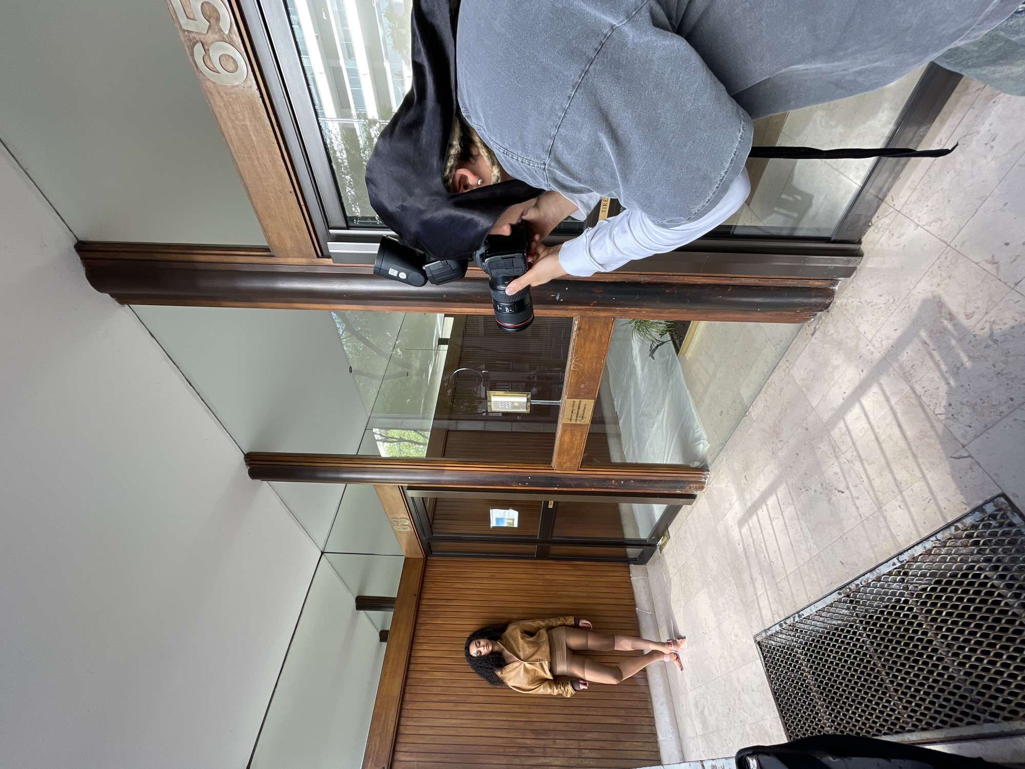 A person in a gray shirt and black hood stands in a doorway, holding a camera and flash, taking a photo of a woman in a tan outfit lying on a wooden surface inside the doorway. The photograph appears to be taken at a building with the number "59" on the facade, likely in Paris, France. The doorway leads to a building with glass walls and wooden framework. The floor outside the doorway is tiled, with a metal grate visible in the lower right corner.