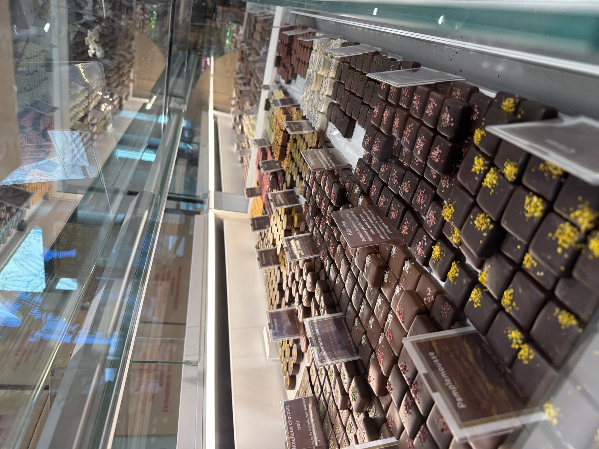 This image captures a close-up view inside a chocolate shop or patisserie in Illkirch-Graffenstaden, France, showcasing an extensive display of artisanal chocolates arranged neatly in a glass refrigerated case. No people are visible.
The primary subjects are rows upon rows of small, square or rectangular chocolates, predominantly dark chocolate, but also some milk and white chocolate varieties. Many of the dark chocolates are adorned with colorful toppings, including pink, yellow, and green sprinkles. These individual chocolates are presented with small, transparent labels indicating their flavors. The setting appears to be a well-lit interior, with reflections on the glass of the display case hinting at overhead lighting.
The visible text on the labels, mostly in French, details the specific flavors and types of chocolates:
* "Moelleux chocolat noir" describes dark soft chocolate candies with a soft ganache filling.
* "Pamplemousse" indicates a grapefruit-flavored chocolate.
* "Charlotte poire" refers to a milk chocolate candy with a pear ganache filling.
* "Mirabelle" denotes a Mirabelle plum-flavored chocolate.
* "Hawaïen" (Hawaiian) describes milk chocolate candies with a coconut ganache filling and pineapple flavor, also accompanied by an English translation.
* Another label partially visible reads "Foret Noire" (Black Forest).
The image emphasizes the variety and detailed craftsmanship of the chocolates, suggesting a high-end confectionery. The clear separation and labeling of each type of chocolate facilitate selection for customers.