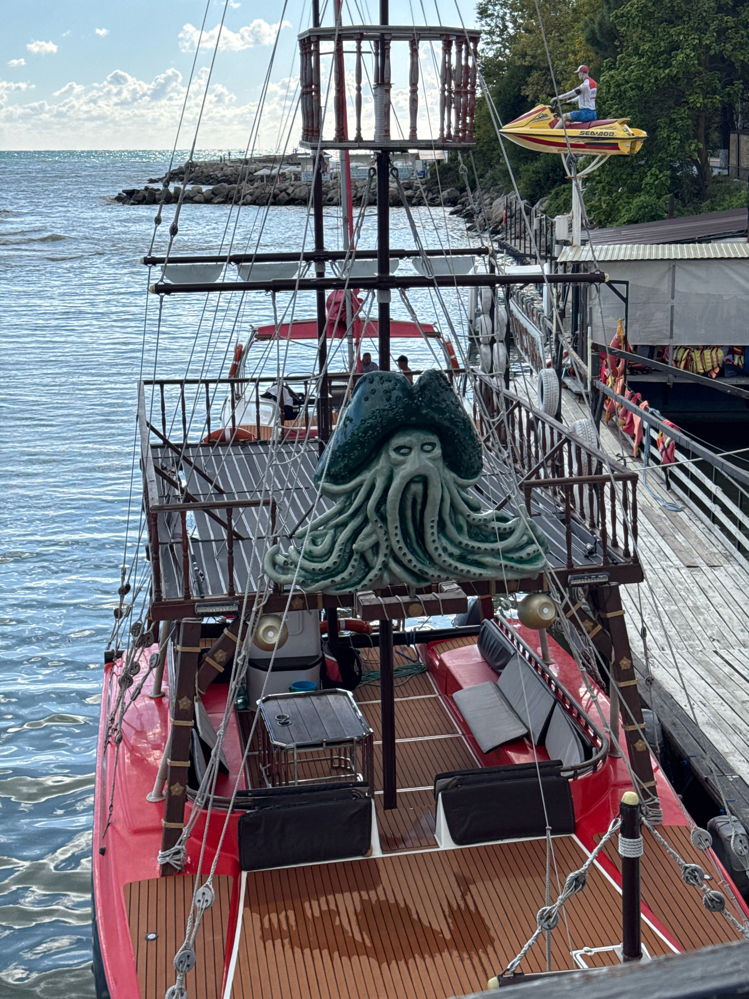 This is a high-angle, medium shot of a red pirate-themed boat docked at a wooden pier in Novomikhaylovsky, Russia. The boat has a prominent, teal-colored Kraken or Davy Jones-like figure with tentacles and a hat at the bow. The deck of the boat is made of wood and features seating areas with gray cushions and black storage compartments. A table is also visible on the deck. Ropes and rigging are strung across the boat, typical of a sailing vessel.
In the background, the sea stretches out to the horizon under a cloudy sky. A rocky shoreline with some structures and beach umbrellas is visible to the left. To the right, a wooden pier runs alongside the boat, with a walkway and what appears to be a building with a dark roof and windows. A yellow jet ski with a person on it is suspended above the pier by a crane-like structure. The overall scene suggests a tourist attraction or recreational area by the sea.