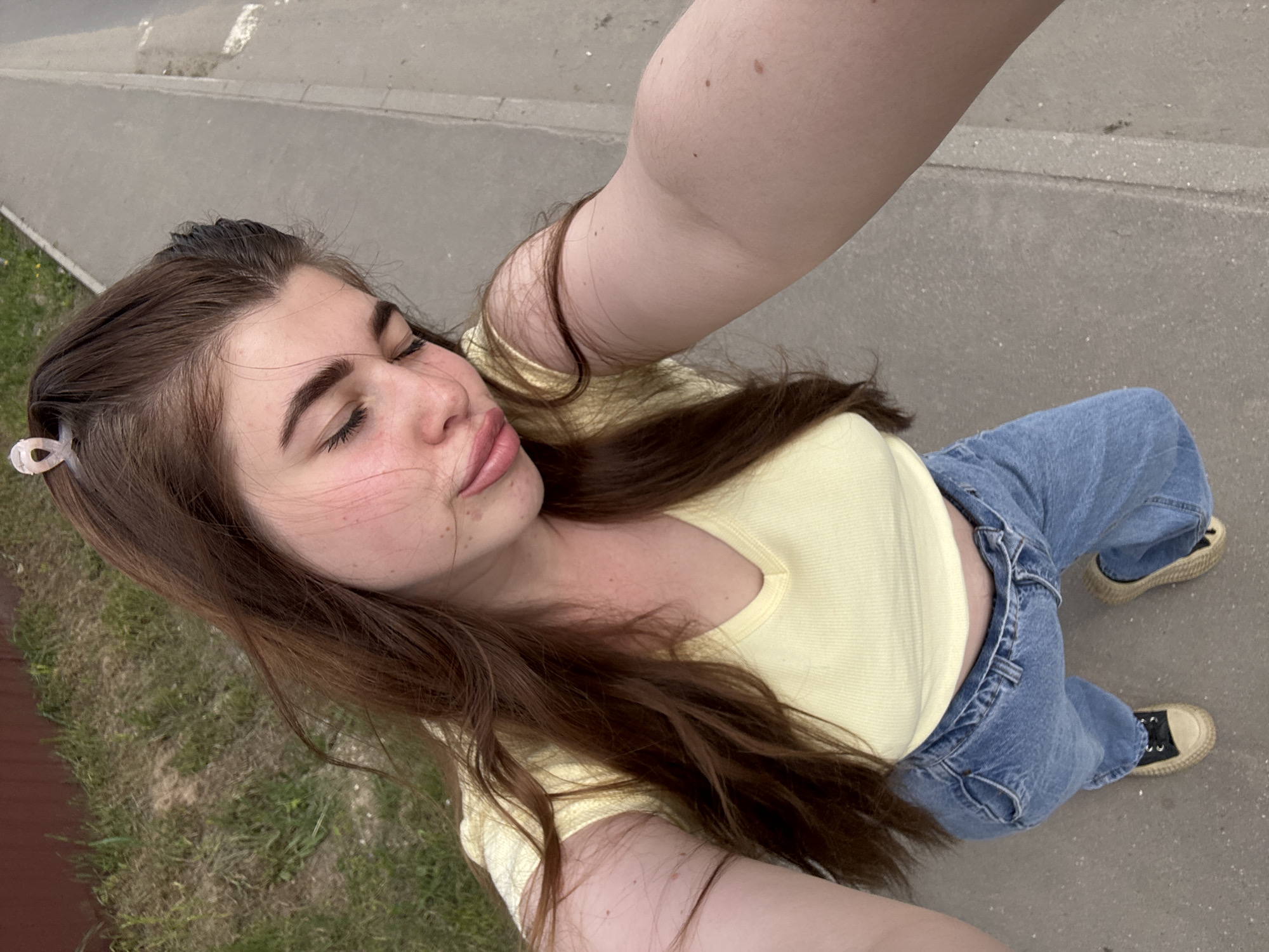 A young woman is taking a selfie outdoors in Posëlok kombinata stroymaterialov-1, Russia. She has long brown hair and is wearing a yellow shirt and blue jeans with dark-colored sneakers. She has her eyes closed and is holding the camera with an arm extended. The background shows a gray paved surface, possibly a sidewalk or road, and some green grass. A part of a wooden fence is visible on the lower left.