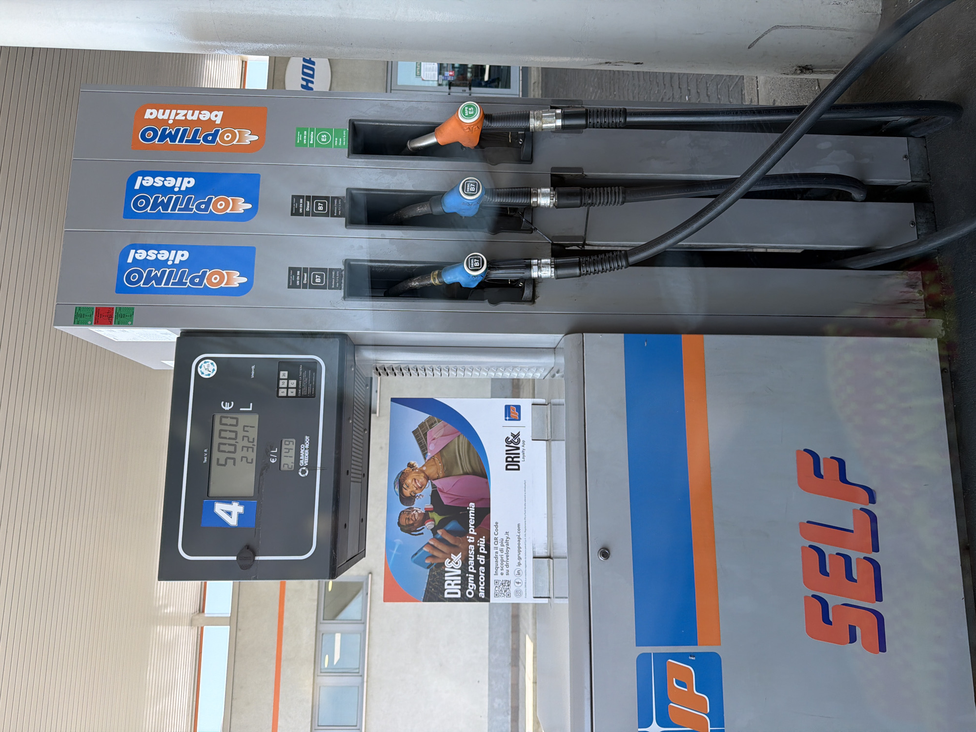 The image shows a self-service gas pump at a gas station in Ferrara, Italy. The pump is a Gilbarco Veeder-Root model and has three nozzles: two for "OPTIMO diesel" and one for "OPTIMO benzina." The display shows a transaction for 50.00 Euros and 23.27 liters at a price of 2.149 Euros per liter. There is a promotional flyer for "DRIVE&" attached to the pump, which encourages customers to scan a QR code to learn more about the loyalty app. The gas station has a white and orange color scheme with "JP" branding on the pump and "SELF" printed on the side. The environment appears to be outdoors with a covered canopy overhead.