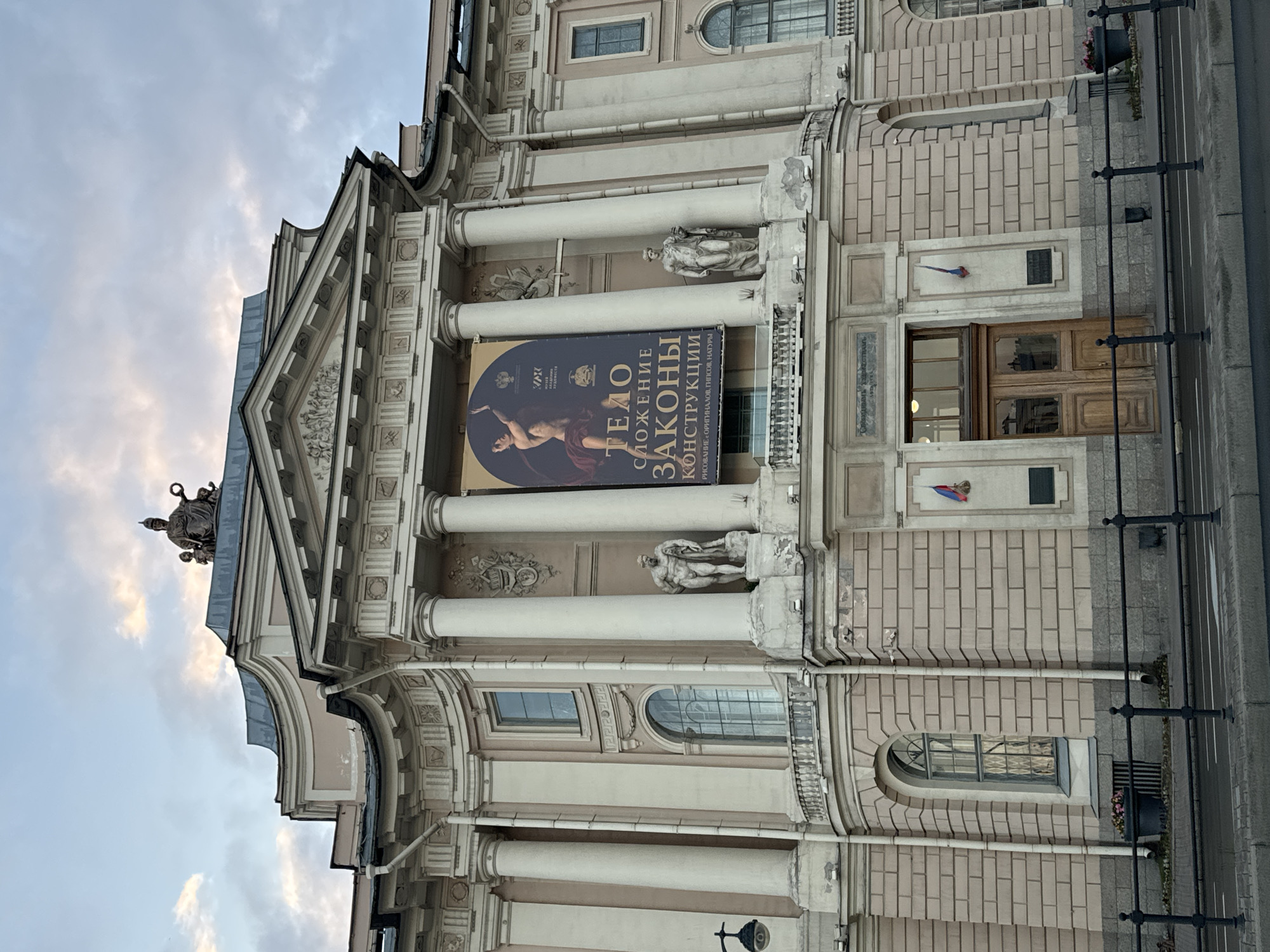 The image shows the facade of a large, classical building in Saint Petersburg, Russia. The building has a light-colored stone exterior with multiple columns, arched windows, and decorative sculptures. A large banner advertising an exhibition titled "Тело. Сложение. Законы. Конструкции" (Body. Composition. Laws. Construction) hangs prominently in the center. The banner features an image of a nude figure and text in Cyrillic. Two sculptures of male figures are positioned on pedestals on either side of the entrance. There is a set of double wooden doors beneath the banner, flanked by flags. The building is set back from a sidewalk and street, with a black metal fence in the foreground. The sky above is cloudy.