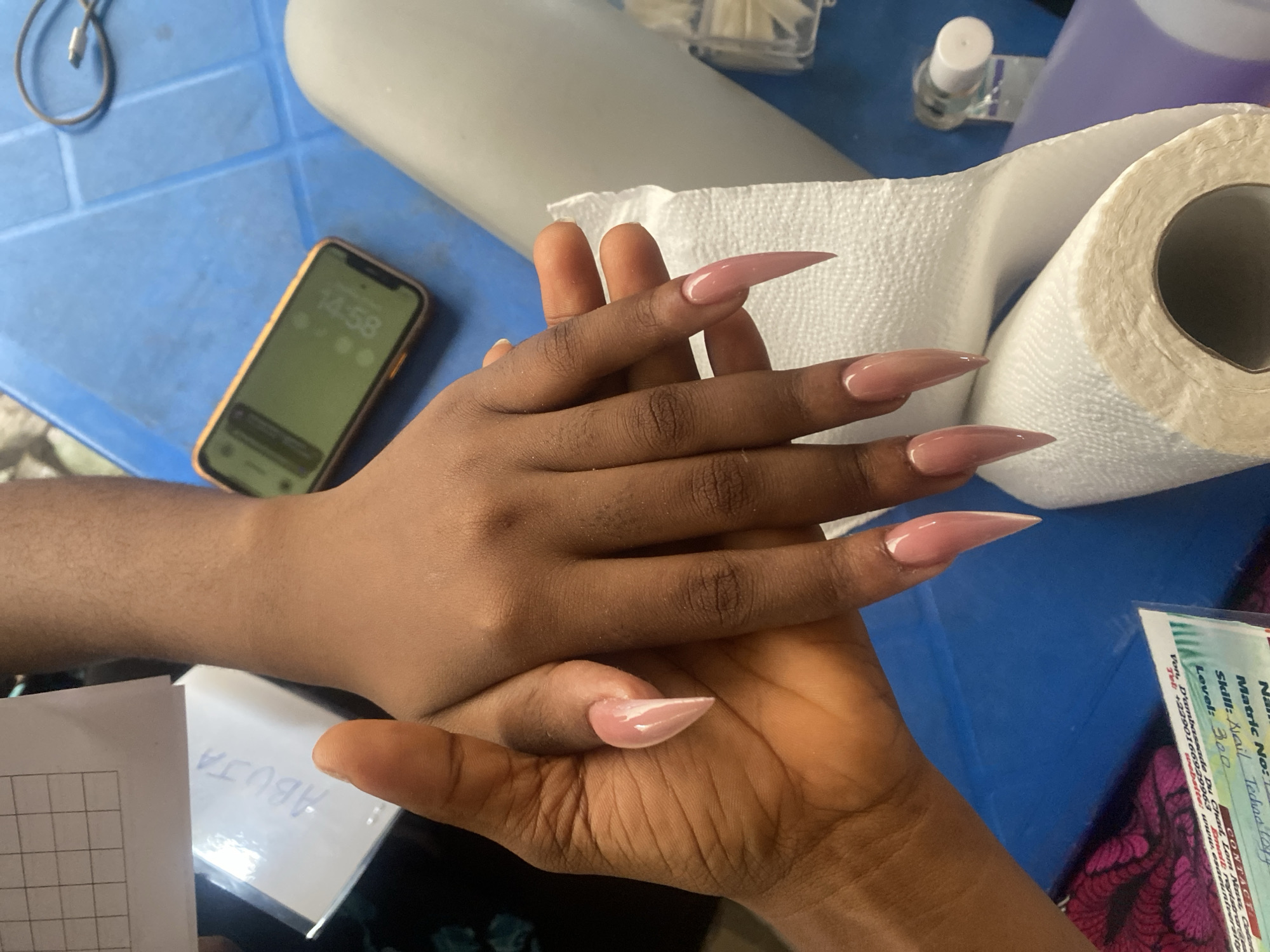 Two individuals are engaged in a nail service, likely in Badagry, Nigeria, with their hands positioned over a blue plastic table. One set of hands, belonging to a client, showcases freshly applied, long, pointed (stiletto or almond shape) acrylic nails in a pale pink or nude shade. The other hand, presumably the nail technician's, gently holds the client's hands, appearing to admire or finalize the work. The warm skin tones of the hands suggest individuals of African descent.
The table is cluttered with various items essential for nail care. A gold-colored iPhone lies unlocked, displaying the time "14:58" on its screen, indicating late afternoon. Beside it, a charging cable is visible. A large roll of white paper towels, clear plastic containers filled with clear nail tips, and a small white bottle (possibly nail glue or polish) are also present. Further details emerge from visible text: a piece of paper partially covered by a hand has "ABUJA" written upside down. Another document or business card provides service details, including "Skill: Nail Technology," "Level: 300," and contact information such as "Von, D'ambassade Du Chad," a Nigerian telephone number (+2390166929963), and an email "registrare". The overall scene depicts a casual yet professional nail artistry session.