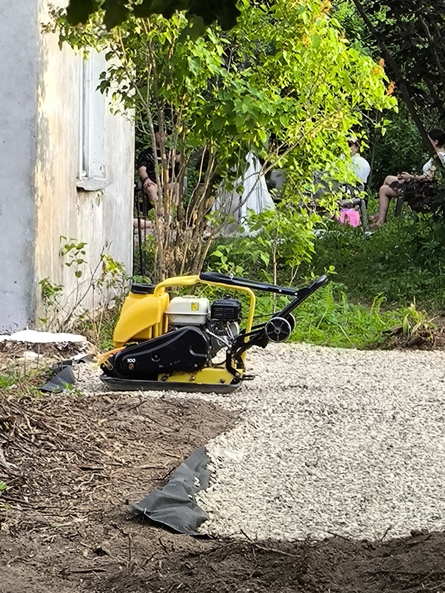 The image depicts an outdoor scene, likely a backyard or garden area, where construction or landscaping work is in progress. In the foreground, a yellow and black plate compactor sits on a bed of light-colored gravel. To the left, a section of a pale, textured wall of a building is visible, partially obscured by green foliage. A black fabric material is laid out on the ground near the gravel. In the background, several people are seated in chairs amidst dense green vegetation. One person wears a white shirt and a cap, another is in a white top and patterned shorts, and another in a white top and dark bottoms. The overall environment is lush with greenery and appears to be a sunny day.