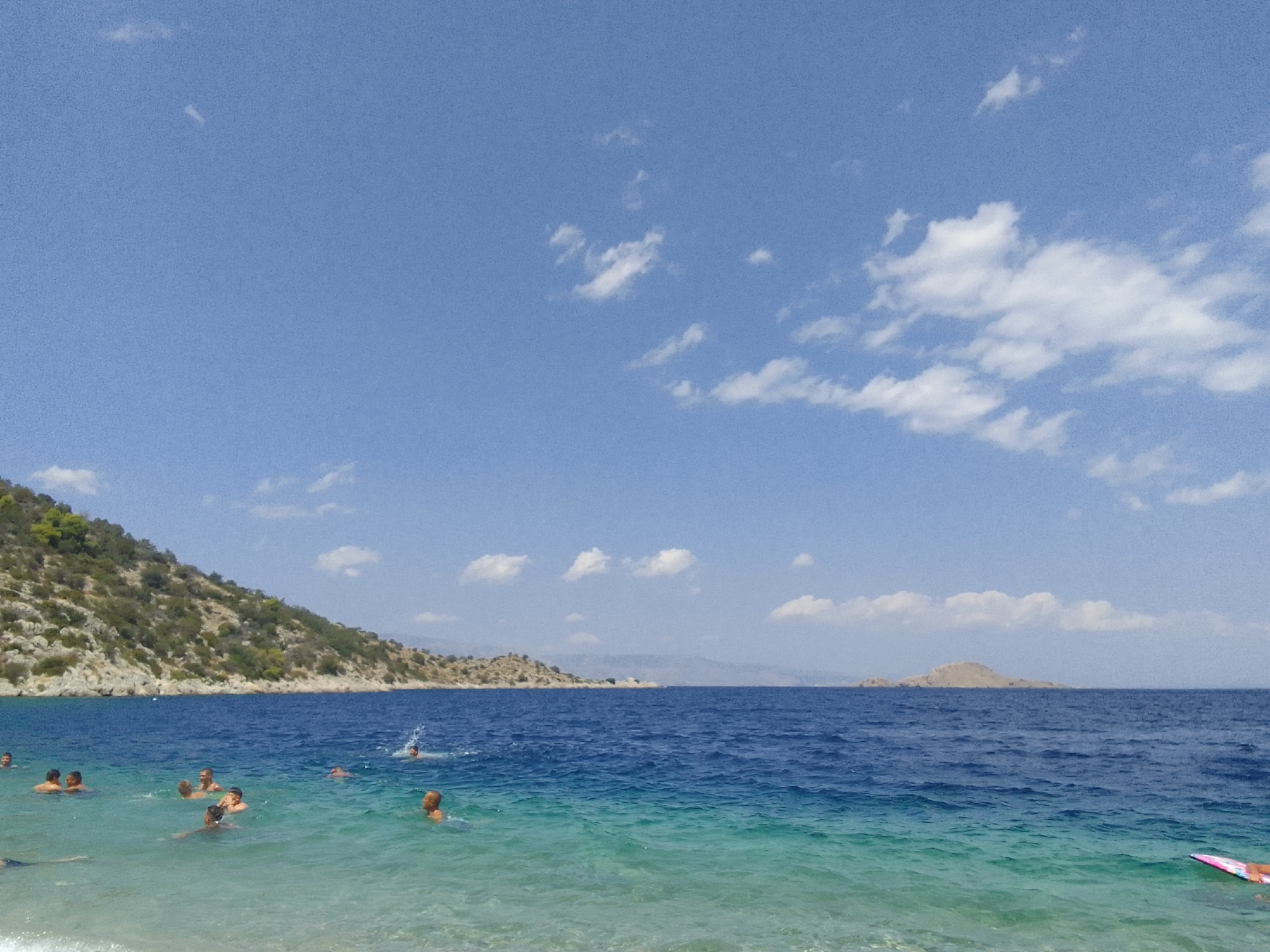An aerial view shows a beach with people swimming and a distant island in the Municipal Unit of Solygeia, Greece. The water is clear and blue, with a rocky coastline in the background. Several people are visible in the water, some swimming and others relaxing. A small island is located in the distance, and clouds are scattered across the sky. The scene is calm and serene, suggesting a pleasant day at the beach.