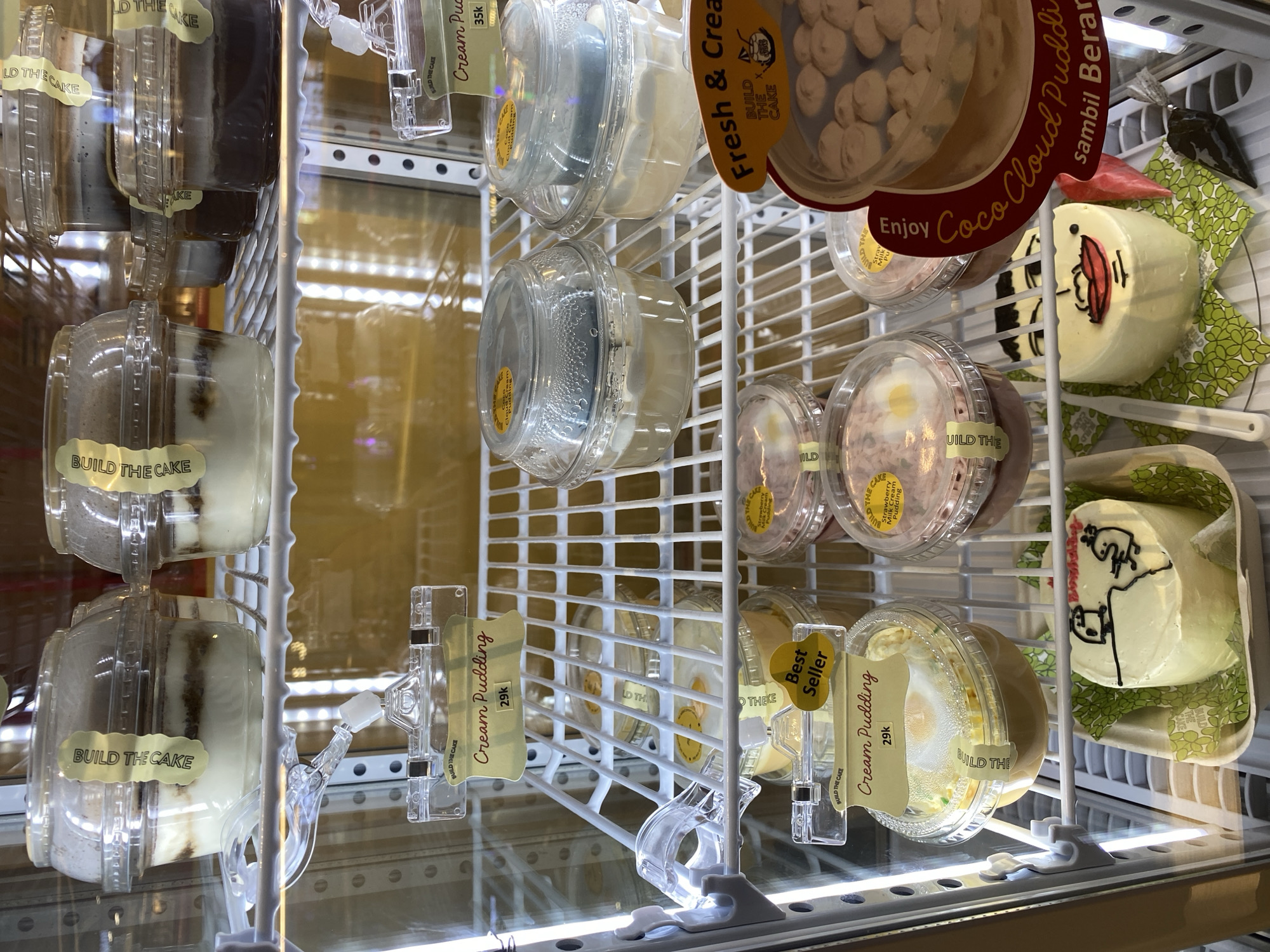 The image shows the interior of a display refrigerator filled with various desserts. The refrigerator shelves are white wire racks, and the items are housed in clear plastic containers. Several desserts are labeled "BUILD THE CAKE" and some have prices like "29k" and "35k". There are layered puddings, what appear to be milk puddings, and a large cake with a face drawn on it. The refrigerator itself appears to be a commercial display unit, likely found in a bakery or dessert shop. The presence of Indonesian currency and a cake with Indonesian text ("sambil Beran") suggests the location is in Indonesia, potentially South Jakarta. The lighting is bright and even, typical of a refrigerated display case. There are no people visible in the image.