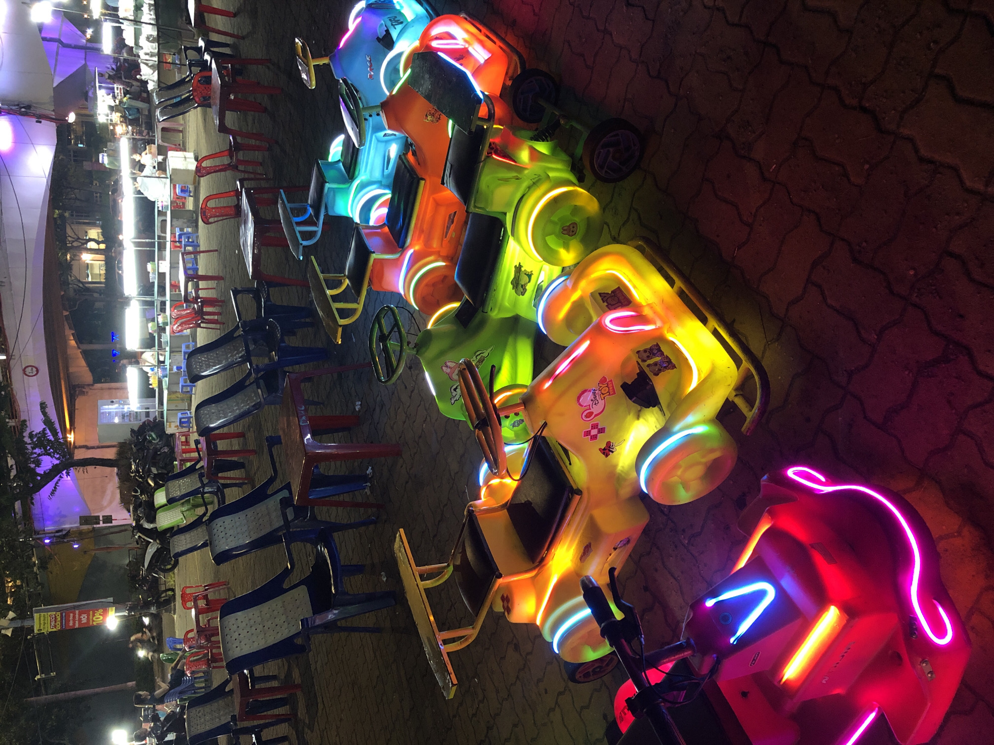 The image captures a vibrant nighttime scene in Đà Nẵng, Vietnam, likely an amusement area or a market. The foreground is dominated by a row of brightly lit, child-sized bumper cars. These cars are in various colors, including yellow, orange, green, and blue, and are adorned with colorful neon lights that outline their shapes and rims. They are parked on a patterned brick pavement.
In the background, numerous plastic chairs and tables are arranged, suggesting a dining or resting area. Some chairs are red, others are blue and gray. Beyond the seating area, there are several stalls or kiosks illuminated by bright fluorescent lights, indicating commercial activity. Motorcycles are parked in the mid-ground. The overall atmosphere is lively and festive, characteristic of an outdoor evening gathering or entertainment spot in Vietnam. The lighting is predominantly artificial, with strong highlights from the neon cars and stall lights, creating a dramatic contrast with the darker surroundings.