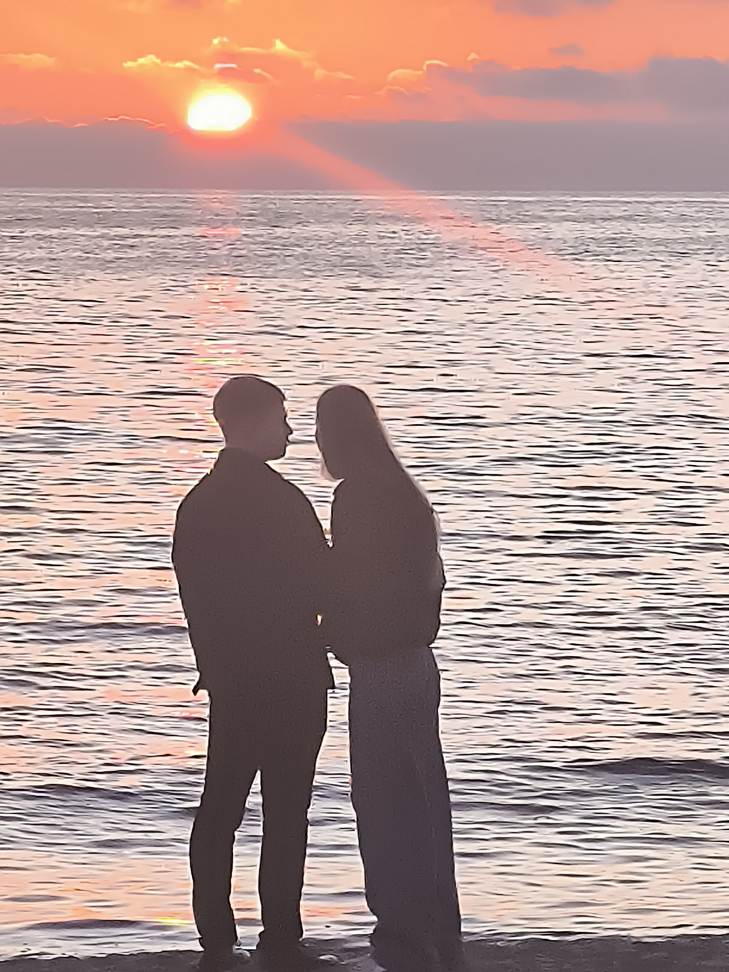 A silhouetted couple stands on a sandy shore, facing each other with the sun setting over the Baltic Sea in Klaipėda, Lithuania. The sun, a bright orb, is partially obscured by soft clouds, casting a warm, orange glow across the sky and reflecting on the rippling water. The couple's figures are dark against the vibrant sunset, their bodies turned towards one another in a tender embrace. The man stands to the left, his arm around the woman. The woman, to the right, has her head tilted towards him. The water stretches to the horizon, its surface a mosaic of pink and orange hues. The scene captures a moment of quiet intimacy against the dramatic backdrop of a natural spectacle.
