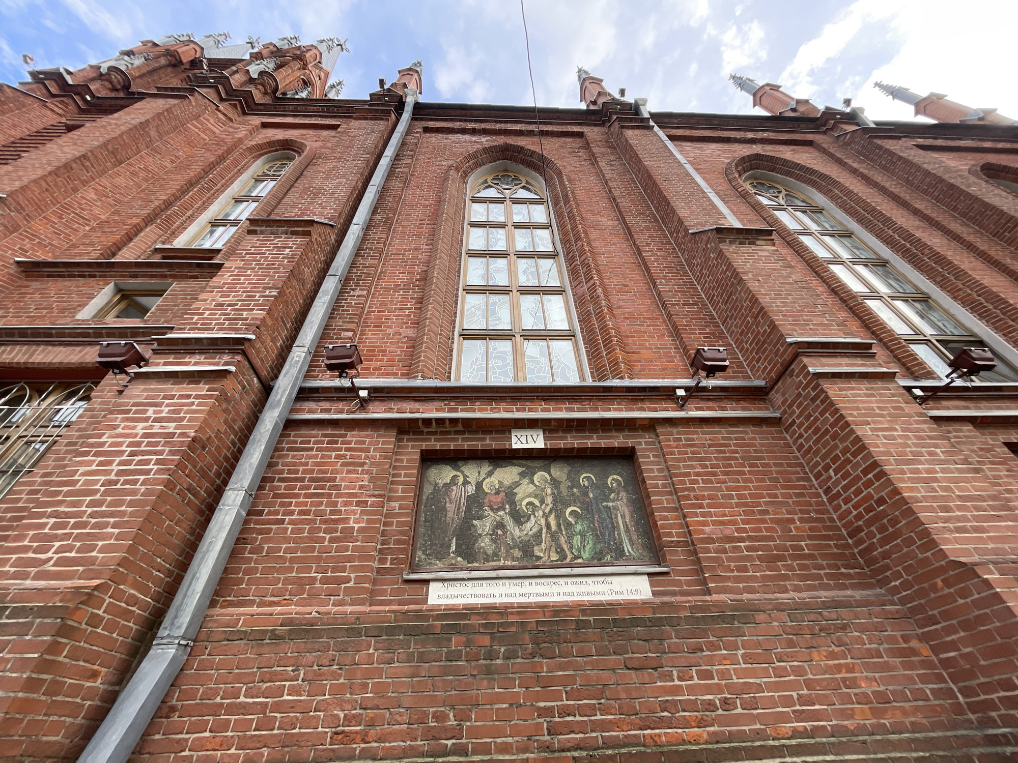 This low-angle, outdoor shot captures a portion of a red brick building with Gothic architectural elements. The building features tall, arched windows with intricate mullions and what appear to be decorative spires along the roofline, suggesting it is a church or a similar historical structure. The brickwork is detailed, with a visible drainpipe running vertically along the wall.
Centrally located on the building is a framed artwork, possibly a mosaic or a painted plaque, depicting a religious scene with multiple figures. Above this artwork, a Roman numeral "XIV" is visible. Below the artwork, there is a white plaque with text in Russian, which appears to be a biblical quote. The sky above is partly cloudy, indicating daytime and fair weather. The overall scene conveys a sense of historical and religious significance, common in older European cities. The text on the plaque reads: "Христос для того и умер, и воскрес, и ожил, чтобы владычествовать и над мертвыми и над живыми (Рим 14:9)", which translates to "Christ died and rose and lived again that he might rule over both the dead and the living (Romans 14:9)." The location context is Moscow, Russia.