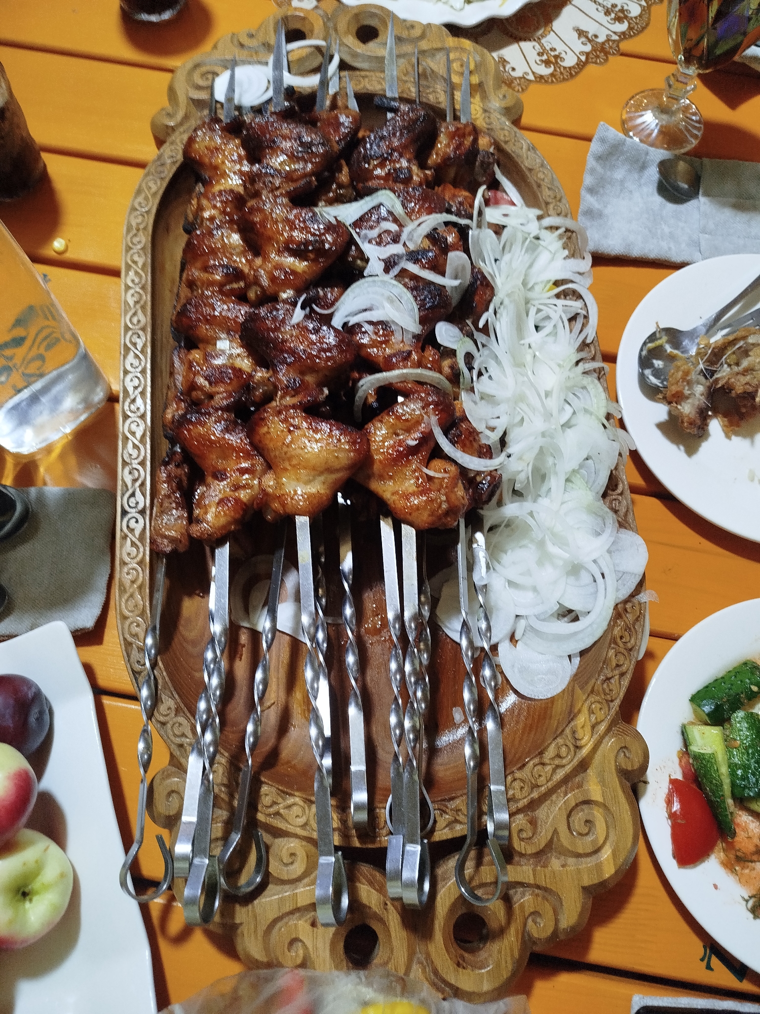A close-up, top-down shot captures a wooden table set with a meal. In the center, a rustic wooden platter holds numerous skewers of grilled meat, interspersed with thinly sliced white onions. The meat appears to be dark brown and glistening. To the right of the platter, a white plate contains what looks like the remains of a dish, with a few pieces of food and cutlery. Further to the right, a small white plate is visible with chopped cucumbers and tomatoes. In the lower left corner, a white rectangular dish holds two apples and a dark plum. The table surface is made of orange-tinted wood planks. In the background, partially visible, are glasses and other dinnerware. The scene is likely from a restaurant or home dining setting in Kabanbay, Kazakhstan, given the food presentation.