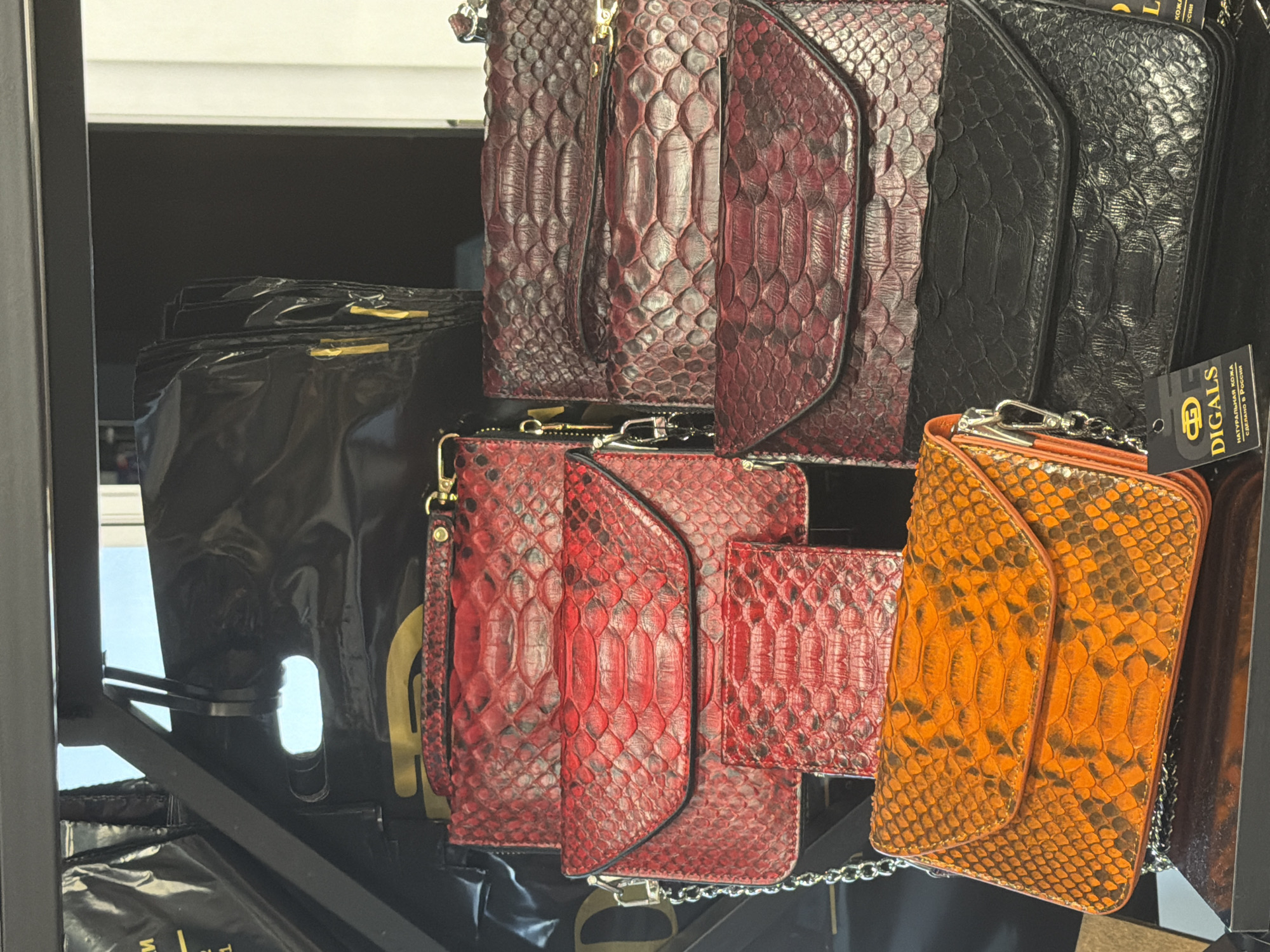 The image shows a display of various python skin handbags and clutches. The handbags are stacked on shelves and are in different colors including red, dark red, orange, and black. Some of the clutches have chain straps. A tag that says "DIGALS" with Russian text is visible on the black clutch. The background appears to be a store or market stall. It is not possible to determine the location in Sochi, Russia, or any specific details about people, actions, or interactions from the provided image.