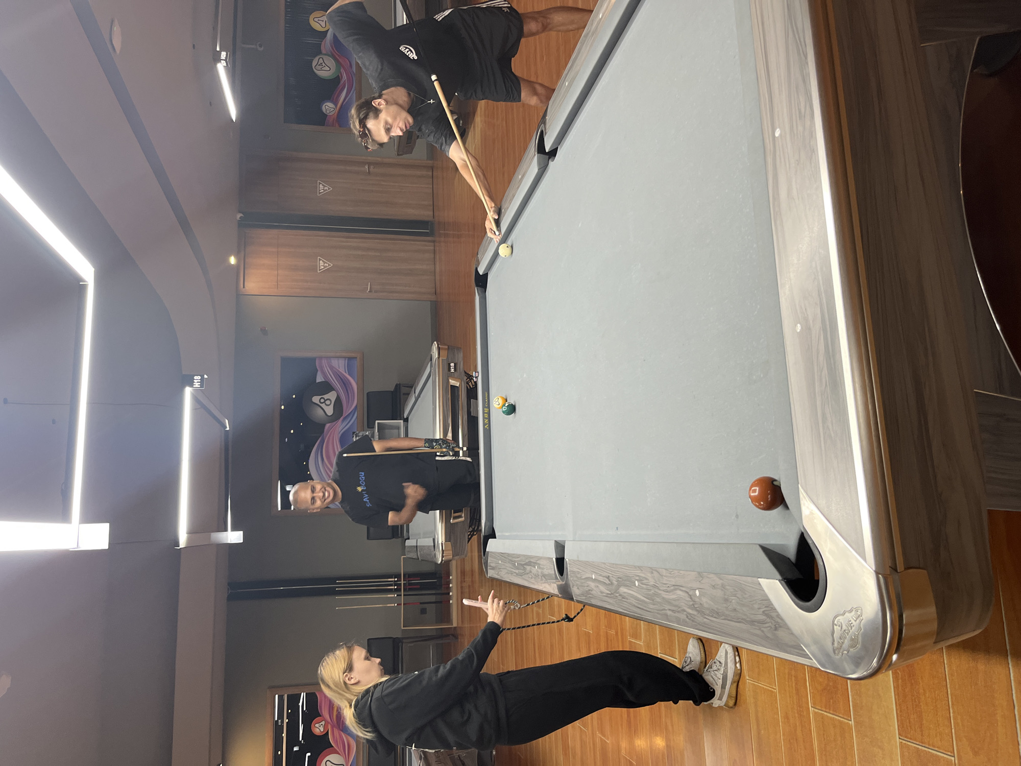 The image depicts three individuals in what appears to be a modern billiard hall in Kuta Selatan, Indonesia. The scene is brightly lit, suggesting daytime or well-illuminated indoor lighting.
In the foreground, a woman with blonde hair is shown from the side, wearing black athletic wear and white sneakers. She is holding a string with her right hand, seemingly engaged in some activity or a game. Her expression is focused.
In the mid-ground, a man with a shaved head is standing near a pool table, wearing a black t-shirt with "SAMY BOGU" printed on the back. He is smiling and looking towards the camera, positioned on the right side of the pool table.
In the background, another man is actively playing pool. He is wearing a black t-shirt and shorts, holding a pool cue and aiming at a ball on the table. His posture indicates concentration. The pool table itself is grey with a decorative wooden frame.
Visible text includes "SAMY BOGU" on the t-shirt of the man in the mid-ground, and decorative text/logos on the pool table pockets and artwork on the walls, which are abstract and colorful. There are also "VIP" logos visible on wooden panels in the background. The flooring is made of polished wooden tiles. The overall atmosphere appears casual and recreational.