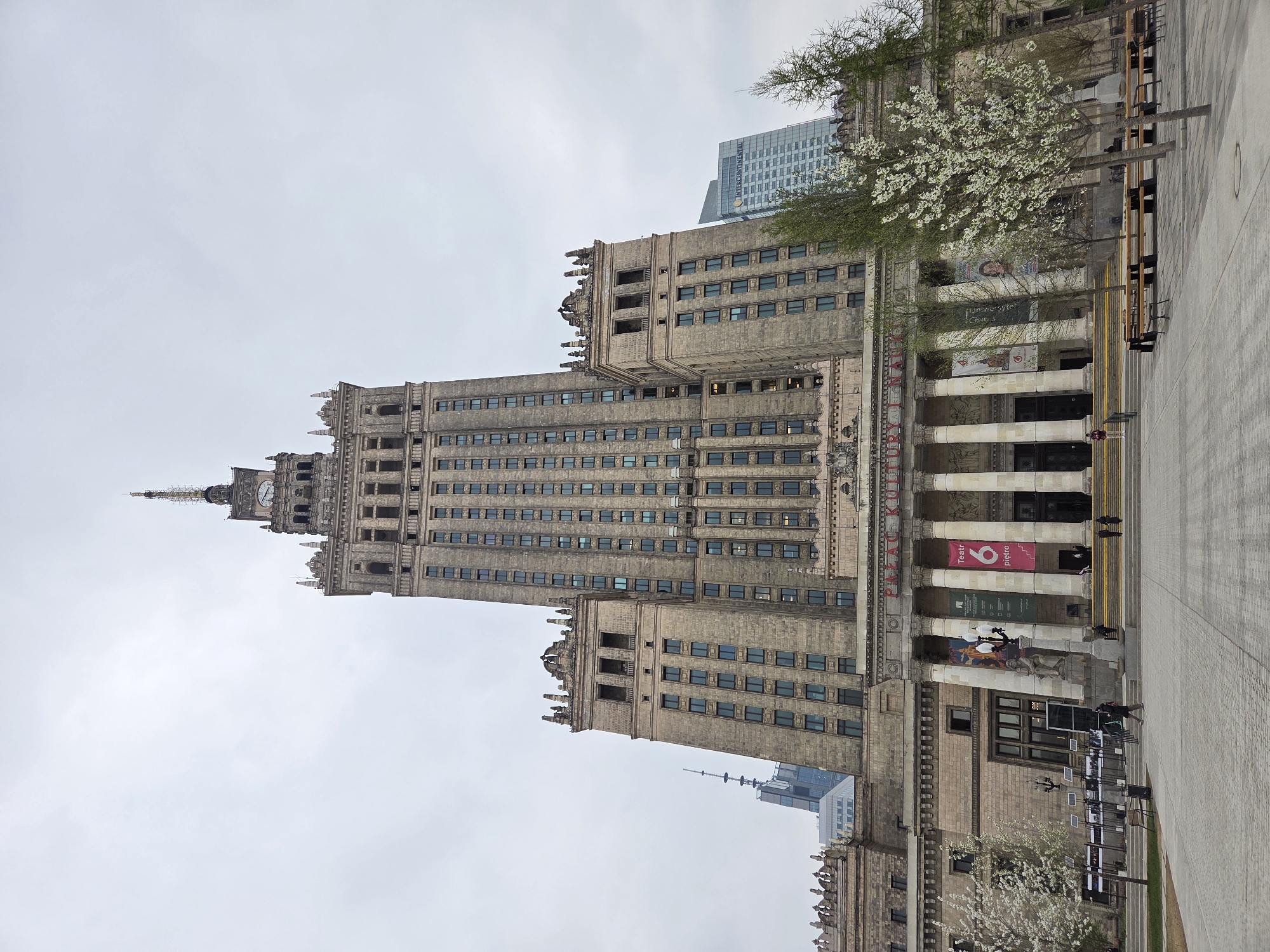 The image shows the Palace of Culture and Science in Warsaw, Poland. The monumental building, a socialist realist structure, dominates the scene. The facade is light brown with numerous windows. A tall spire rises from the top. In the foreground, a wide walkway with a few people is visible. Some posters and banners are displayed on the colonnades of the building. Blooming trees are visible near the base of the building. The sky is overcast. Another, modern building can be seen in the background.