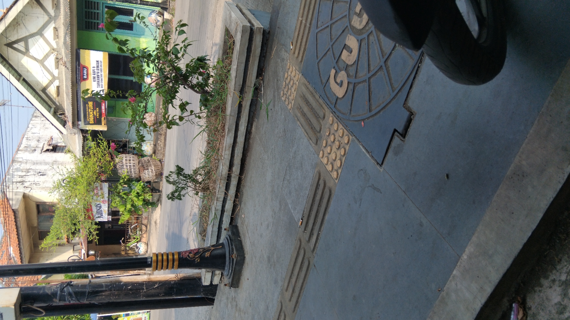 The image depicts a street-level view of a sidewalk and road in Batang, Indonesia, likely in front of a small commercial establishment. On the sidewalk, there is a concrete planter box containing sparse vegetation and dry grass. In the foreground, textured paving blocks for the visually impaired are visible. Adjacent to these blocks is a dark manhole cover with circular and geometric patterns. A motorcycle's tire is partially visible in the lower right corner. The background shows a building with a light-colored facade and a tiled roof, a street with passing vehicles, and other buildings with signage. The overall scene suggests an urban environment on a bright day.