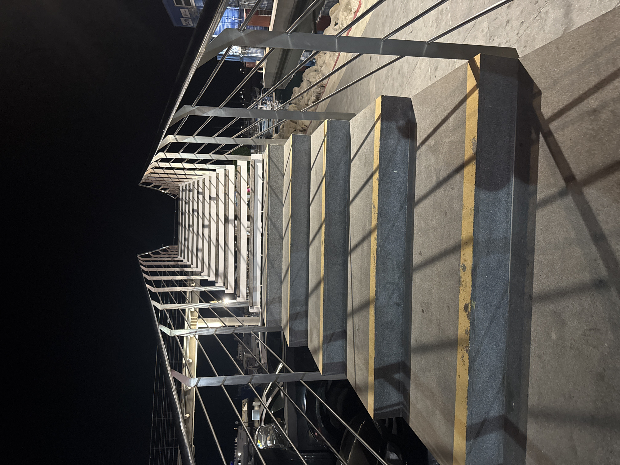 The image shows a modern pedestrian overpass at night, likely located in Limassol, Cyprus, given the context of the query. The overpass features a concrete pathway leading up a series of wide, gray steps, each marked with a yellow stripe along its edge. The steps are flanked by sleek, metallic railings with multiple horizontal bars, supported by vertical posts. Lights illuminate the underside of the steps and the railing structure, casting long shadows on the pathway. In the background, the dark night sky is visible, with faint lights suggesting urban surroundings. There are no people visible in the image. The lower section of the overpass appears to be adjacent to a road with parked vehicles, and further beyond, a body of water with boats and rocky shoreline can be seen, hinting at a coastal or marina area. The overall scene is clean and contemporary.