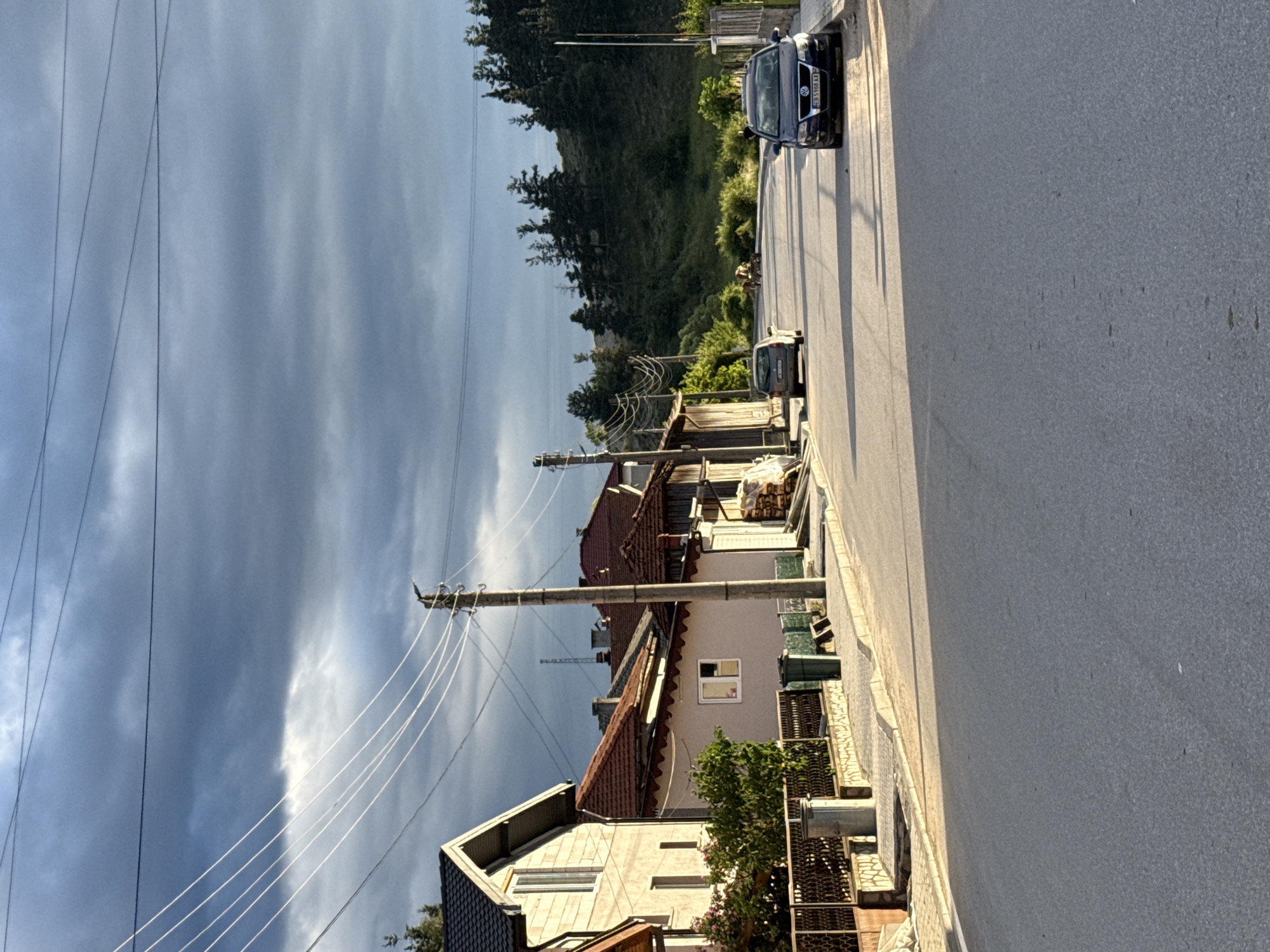 This is a photograph taken in Barutin, Bulgaria, showing a street scene. The image features a paved road with a sidewalk on the left side. Several houses with red tiled roofs are visible along the sidewalk. Electrical poles and wires run across the scene. Two cars are parked on the right side of the road. In the background, there is a wooded hillside with trees. The sky is overcast with dark clouds. The overall impression is of a rural or semi-rural residential area.