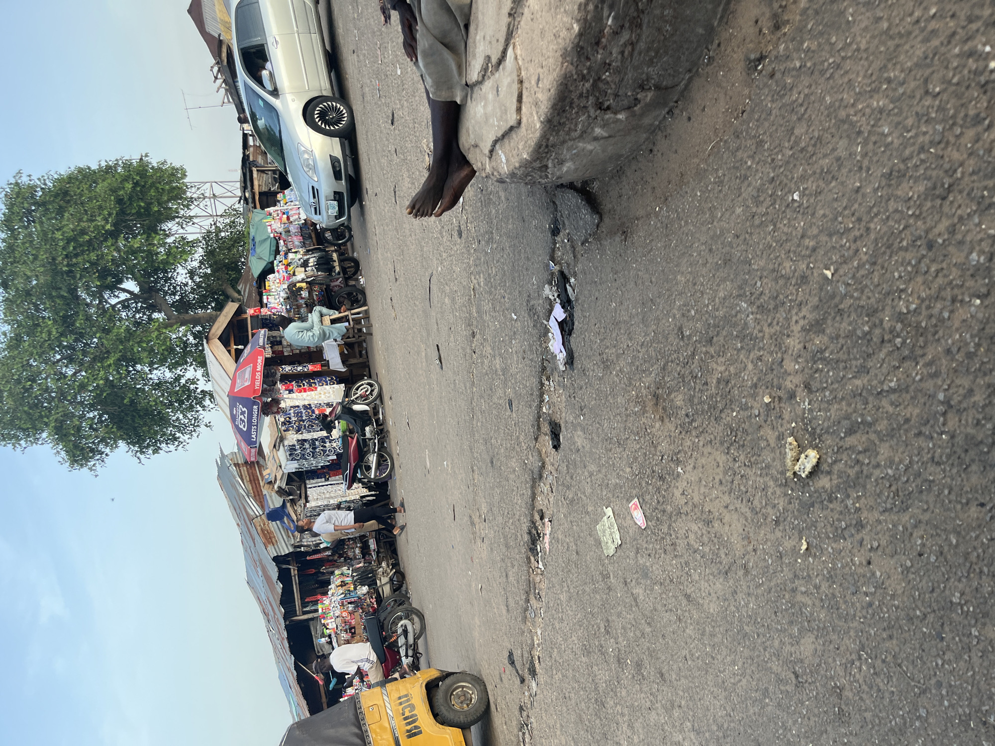 The image depicts a street scene in an urban environment, likely a market area, under a clear sky. In the foreground, a section of paved road shows signs of wear and tear with visible potholes and scattered debris, including paper and what appears to be food waste. A large, concrete curb occupies the right side of the frame, with a person's bare feet resting on its edge. The person is wearing light-colored pants.
To the left, a yellow, three-wheeled vehicle, possibly a tuk-tuk, is parked. Behind it, a red motorcycle is positioned. Further into the scene, a row of stalls with corrugated metal roofs are set up along the street. These stalls display various goods, including textiles with patterned designs, and other merchandise. Several individuals are visible around the stalls. One person, wearing a white t-shirt and dark pants, is walking towards the stalls. Another person, dressed in a light blue garment, is seated on a stool in front of a stall, appearing to be a vendor or shopper. A silver sedan is driving on the road in the mid-ground, with a person visible inside. A large tree with dense foliage is situated behind the stalls, providing shade. In the background, more structures and possibly utility poles are discernible, suggesting a built-up area. The lighting indicates it is either early morning or late afternoon.