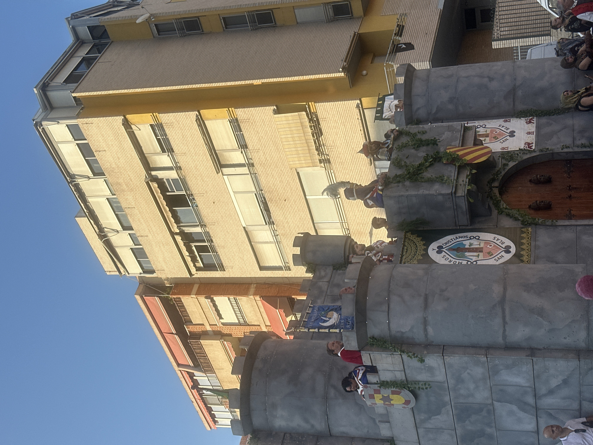 A recreation of a castle structure, adorned with ivy, is positioned in front of a building with yellow and beige brickwork, and red awnings. Several people dressed in historical costumes are visible on top of the castle structure, some holding banners and wearing helmets. One banner on the castle structure displays "MOROS Y CRISTIANOS SAN BLAS." The scene appears to be set on a street in Alicante, Spain, as indicated by the banner referencing "San Blas," a common patron saint's festival location in Spanish cities, and the architectural style of the building which is consistent with Mediterranean urban environments. The sky is clear and blue, suggesting a daytime setting.