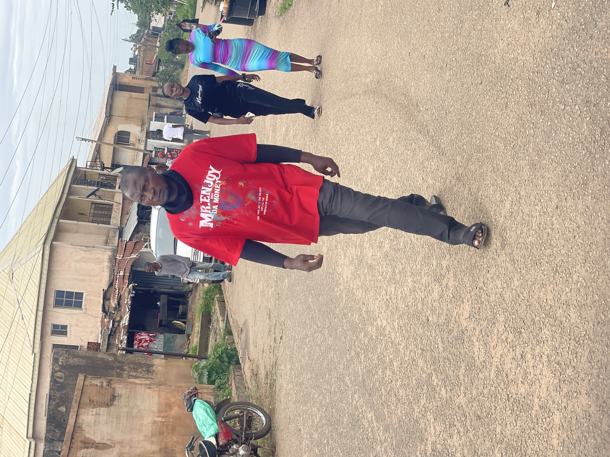 The image depicts a street scene in Jos, Nigeria, under an overcast sky. In the foreground, a man walks towards the camera, wearing a red t-shirt with graphics and text, black pants, and sandals. To his right, a woman in a black t-shirt and pants, and another woman in a colorful, horizontally striped dress walk away from the camera. Behind them, several other people are visible in the distance. The street is paved with asphalt, and on the left side, a motorcycle is parked. The background features a row of buildings with corrugated metal roofs, typical of urban architecture in the area. Some buildings appear to be shops or residences. The overall impression is a snapshot of daily life on a street in Jos.