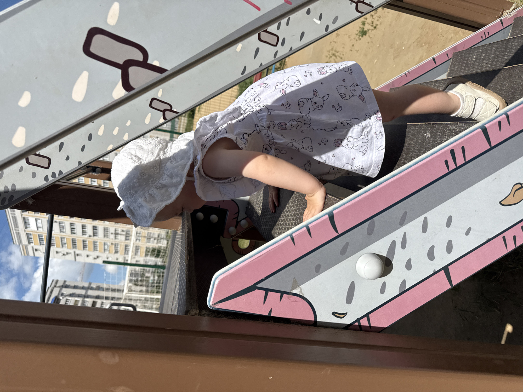 A young child wearing a white sun hat and a white dress with a bunny pattern is climbing on a playground structure. The child is facing away from the camera and towards a metal slide with cartoonish drawings. In the background, tall residential buildings are visible, characteristic of the city of Vladimir, Russia. The playground appears to be made of metal and wood, and the ground is covered in sand. The sky is blue with some clouds.
