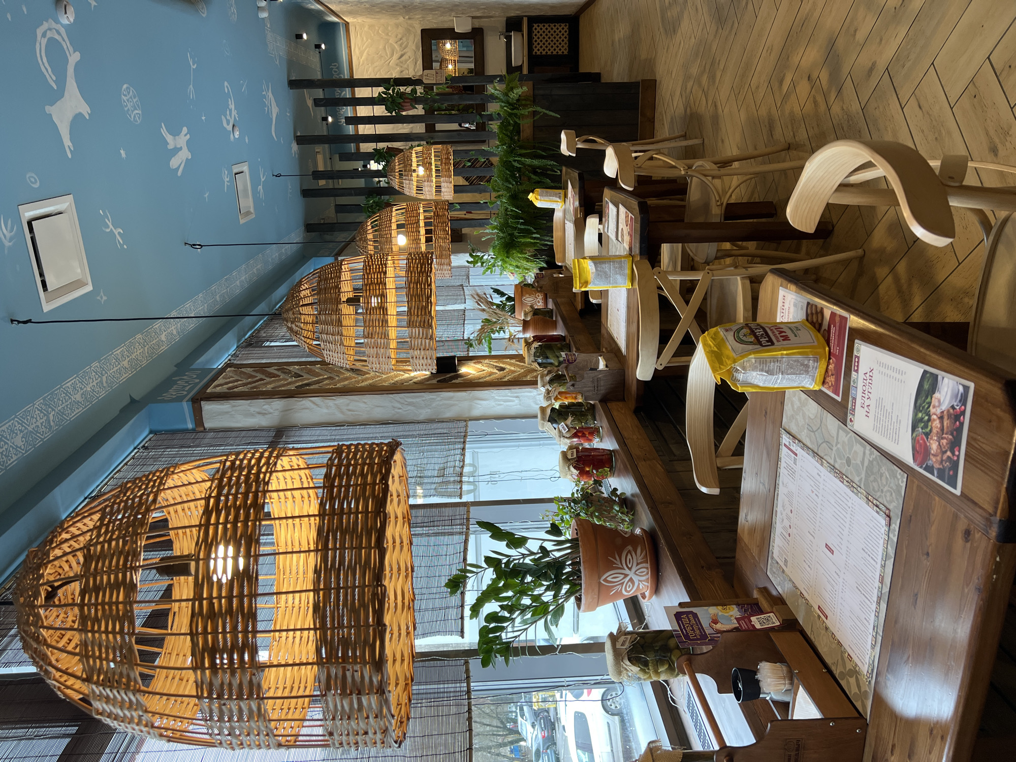 This image captures the interior of what appears to be a Georgian restaurant or cafe, distinguished by its unique decor. The scene is devoid of people, suggesting it might be closed or captured before opening hours.
The most striking feature is the light blue ceiling, adorned with white, primitive-style drawings reminiscent of cave art, depicting animals like deer or goats and human-like figures. A decorative white pattern borders the top of the walls. Hanging prominently from the ceiling are three large, elongated pendant lights made of woven wicker, casting a warm glow.
The dining area features long, dark wooden tables and light wooden chairs, some with curved backs and others appearing to be foldable. Several tables are set with menus in Cyrillic script, displaying various dishes including an image of shashlik (skewered meat). On the tables, there are small condiment holders and a yellow bag labeled "REZEPB MYKI," which is likely a brand of flour (Рецепт Муки - Flour Recipe).
Along a shelf running parallel to a large window, various potted plants, including a fern-like plant and a ZZ plant in a decorated terracotta pot, add a touch of greenery. Jars of preserved foods like pickles and what appear to be pickled tomatoes are also displayed, some labeled in Cyrillic, for example, "ПО-ГРУЗИНСКИ" (Georgian style).
Through the large window, an urban street scene with cars and trees is visible, indicating a daytime setting, possibly overcast or late afternoon. The overall ambiance is rustic and inviting, consistent with a traditional Georgian dining experience.