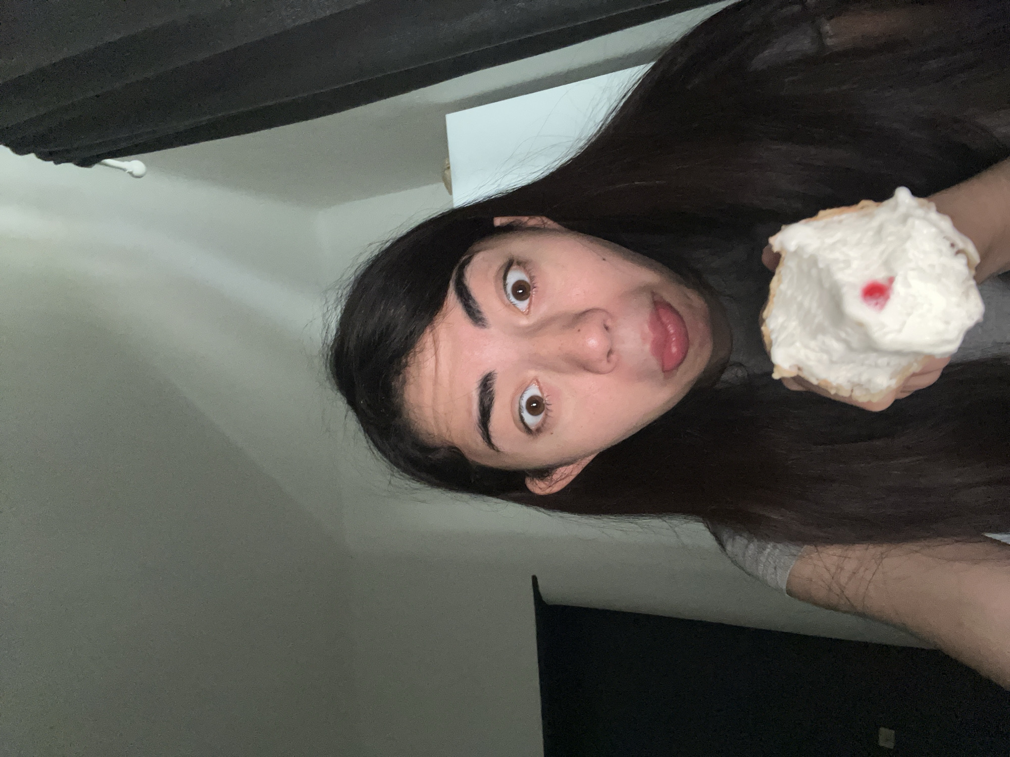 A young woman with long, dark hair is holding a partially eaten pastry with white frosting and a red dollop. She is looking directly at the camera with wide eyes and her tongue sticking out slightly. She is wearing a grey t-shirt. The background is dimly lit, with dark curtains to the right and a white wall to the left. The image was taken in a domestic setting.