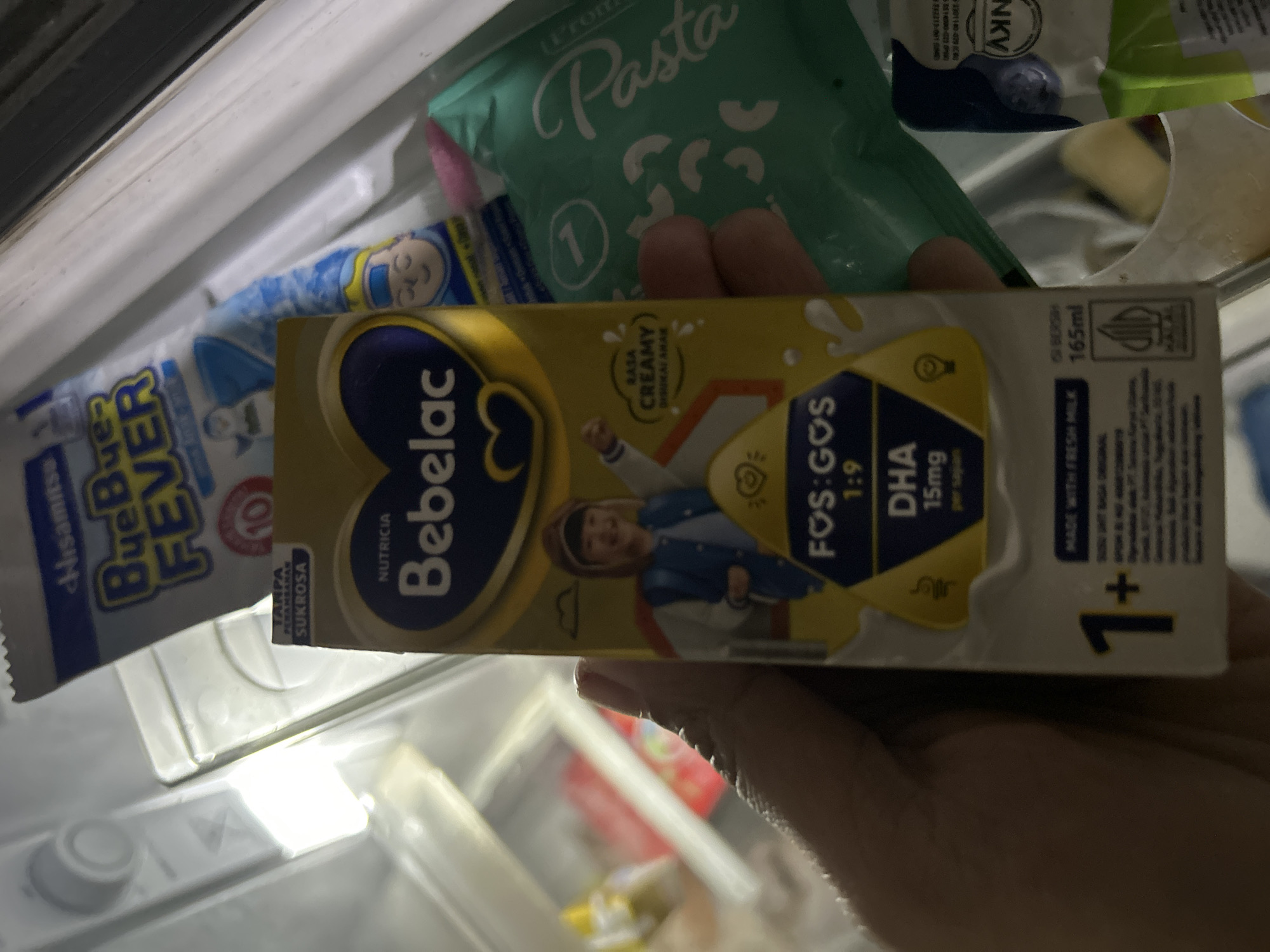 The image shows a hand holding a carton of Bebela milk, with other items in the background, possibly inside a refrigerator. The Bebela milk carton is yellow and blue, with a picture of a child on it. It also has text in Indonesian, including "NUTRICIA," "BebelaC," "FOS:GOS 1:9," and "DHA 15mg per saji." In the background, there are other products, including a blue and white packet labeled "Bye-Bye Fever," and a green packet labeled "Pasta." The overall environment appears to be indoors and possibly a domestic setting, given the presence of food items. The image does not contain any information that would allow for identification of the location within Depok, Indonesia.