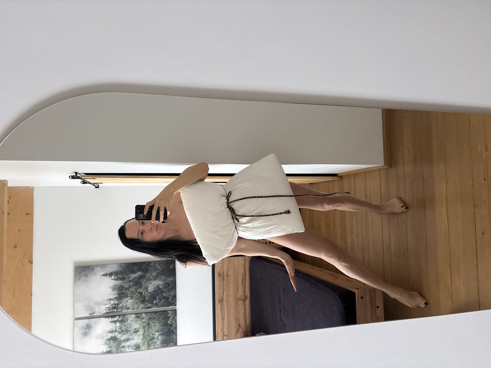 The image depicts a reflection in a rounded mirror capturing a woman holding a pillow. She holds a mobile phone in her right hand, presumably taking the photo. Her long dark hair contrasts with her fair skin. A white pillow tied with a patterned cord partially obscures her body. The background includes a bed with dark bedding, a wooden headboard, and wood flooring. A painting of a forest scene is visible on the wall. The precise location in Chaikovsky, Russia, is not discernible from the image.
