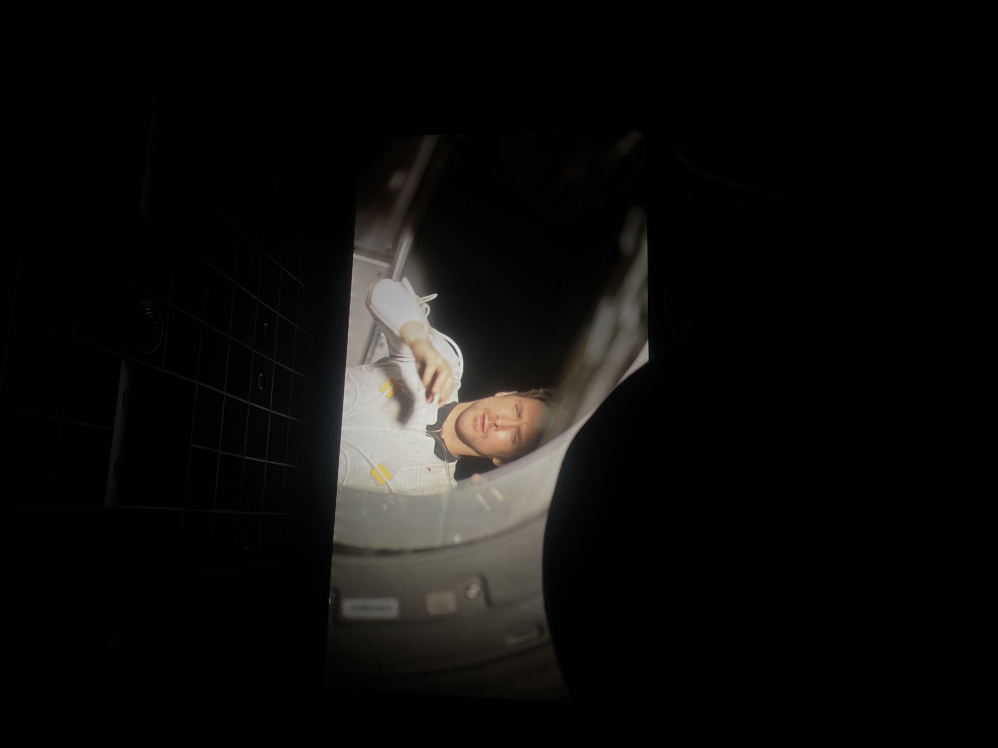 An astronaut is seen inside of a cylindrical space, presumably a spacecraft or simulator. The astronaut is wearing a white spacesuit with yellow accents. The person's face is visible and the lighting is focused on the astronaut's face and body. The background shows metallic surfaces and structural elements, typical of a spacecraft interior. The photograph appears to have been taken indoors, possibly in a space training facility or a museum exhibit. The location details (Krasnoyarsk, Russia) are not visible in the image.