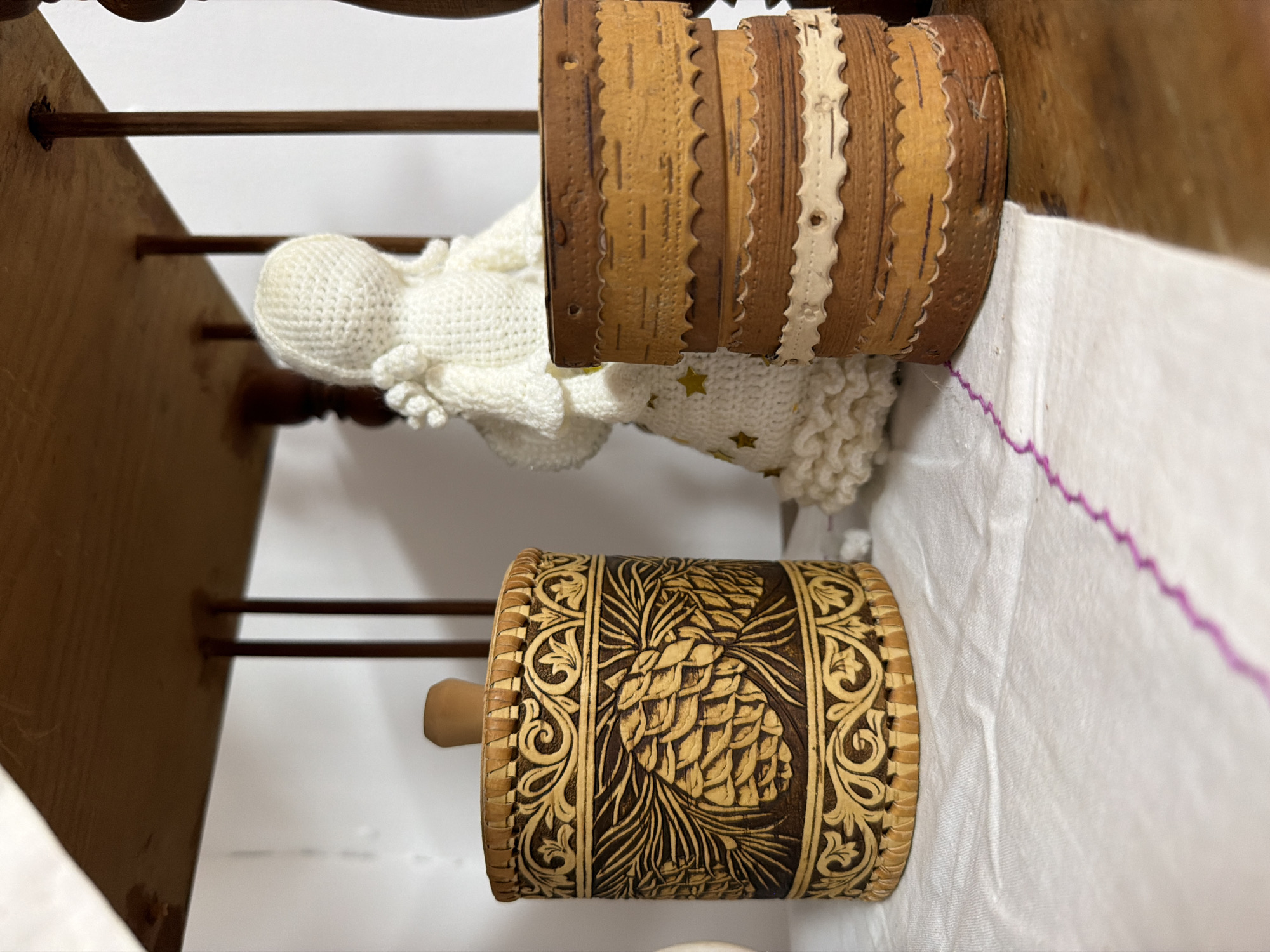 The image shows a close-up view of a wooden shelf in a room. On the shelf are two birch bark containers and a crocheted doll. The container on the left is larger and cylindrical, with intricate carvings of pinecones and swirling patterns. It has a wooden knob on top. To the right of it, there are two smaller stacked birch bark containers with decorative scalloped edges and ribbon accents. Behind these objects, a white crocheted doll with a conical head and ruffled clothing is partially visible. The shelf itself appears to be made of dark wood, and the background is mostly a plain white wall with some indistinct wooden elements on the right. The items are placed on a white cloth with a visible pink embroidered edge. The image does not provide any information about the city of Ust'-Kabyrza, Russia, or any specific context related to it.