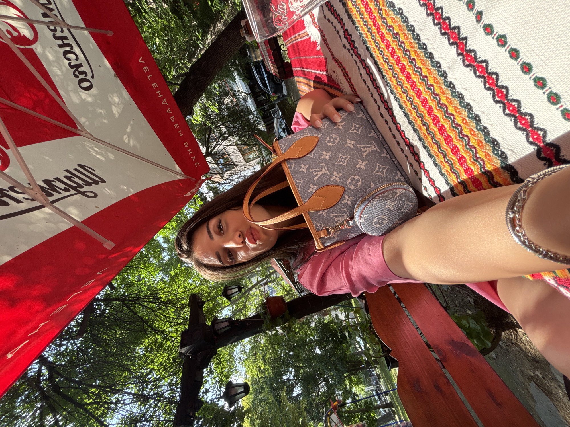 The image, rotated counter-clockwise, depicts a young woman posing for a selfie at an outdoor cafe or restaurant. She is seated, with her face, dark hair, and part of her torso visible as she looks towards the camera. She wears a pink top. She holds a gray handbag featuring a distinctive monogram pattern (resembling Louis Vuitton) with tan leather accents, and a small circular pouch attached. A silver chain bracelet is visible on her wrist or arm.
Adjacent to her, a colorful, horizontally striped textile with geometric patterns, possibly a traditional Bulgarian rug or tablecloth, covers a table surface. A wooden bench or chair in a reddish-brown hue is partially visible below her.
Above, a large red and white striped umbrella provides shade. Cyrillic text is visible on the umbrella: "VELEHAYDEH BKAC" (likely "Велехаудех ВКУС", suggesting a brand like "Velhada Taste" in Bulgarian) along one edge, and partially "МЪШЧКО" (possibly "Майсторско," meaning "Masterful" or "Artisan").
The background is dominated by lush green trees, indicative of a sunny, outdoor environment with clear weather. Sunlight creates distinct shadows. Visible within the foliage are decorative outdoor lamps or lanterns mounted on a wooden beam. The scene suggests a relaxed atmosphere in an al fresco setting, consistent with a public space in Plovdiv, Bulgaria.