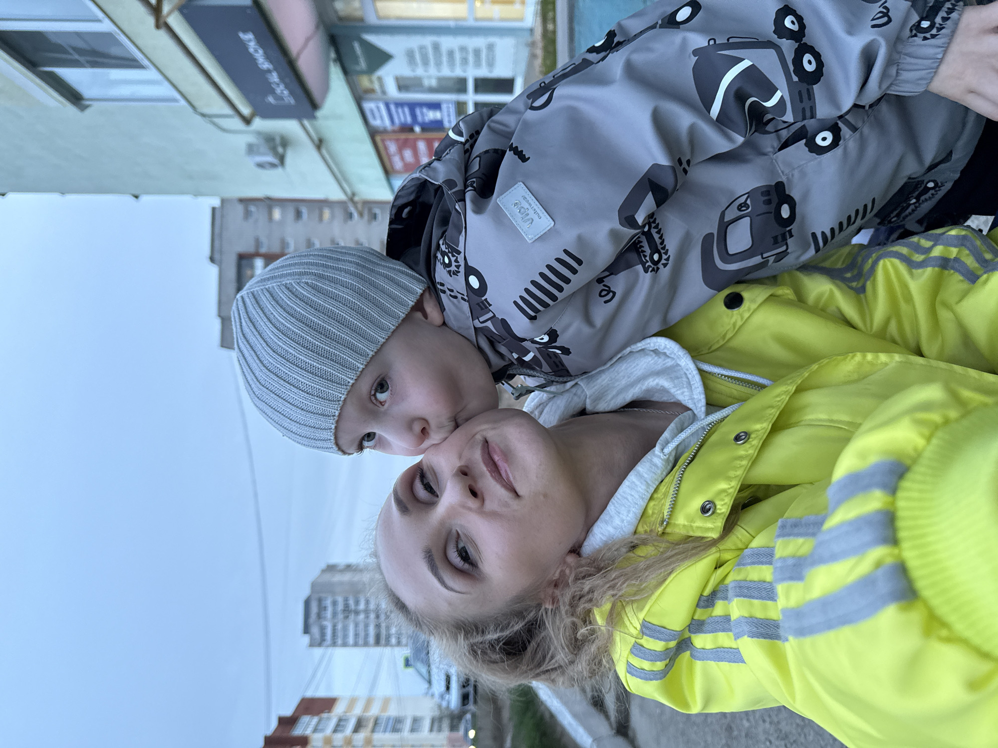 A selfie of a woman and a young child is taken outdoors with buildings in the background. The woman has blonde hair and is wearing a bright yellow jacket with reflective stripes on the sleeve. The child, wearing a gray beanie and a gray jacket patterned with drawings of trucks, is kissing the woman's cheek.
The location is likely the city of Chaikovsky, Russia, suggested by the architecture and general urban environment. Buildings of varying heights are visible in the background under a cloudy sky. The buildings range from low-rise to high-rise, indicative of a modern urban area.