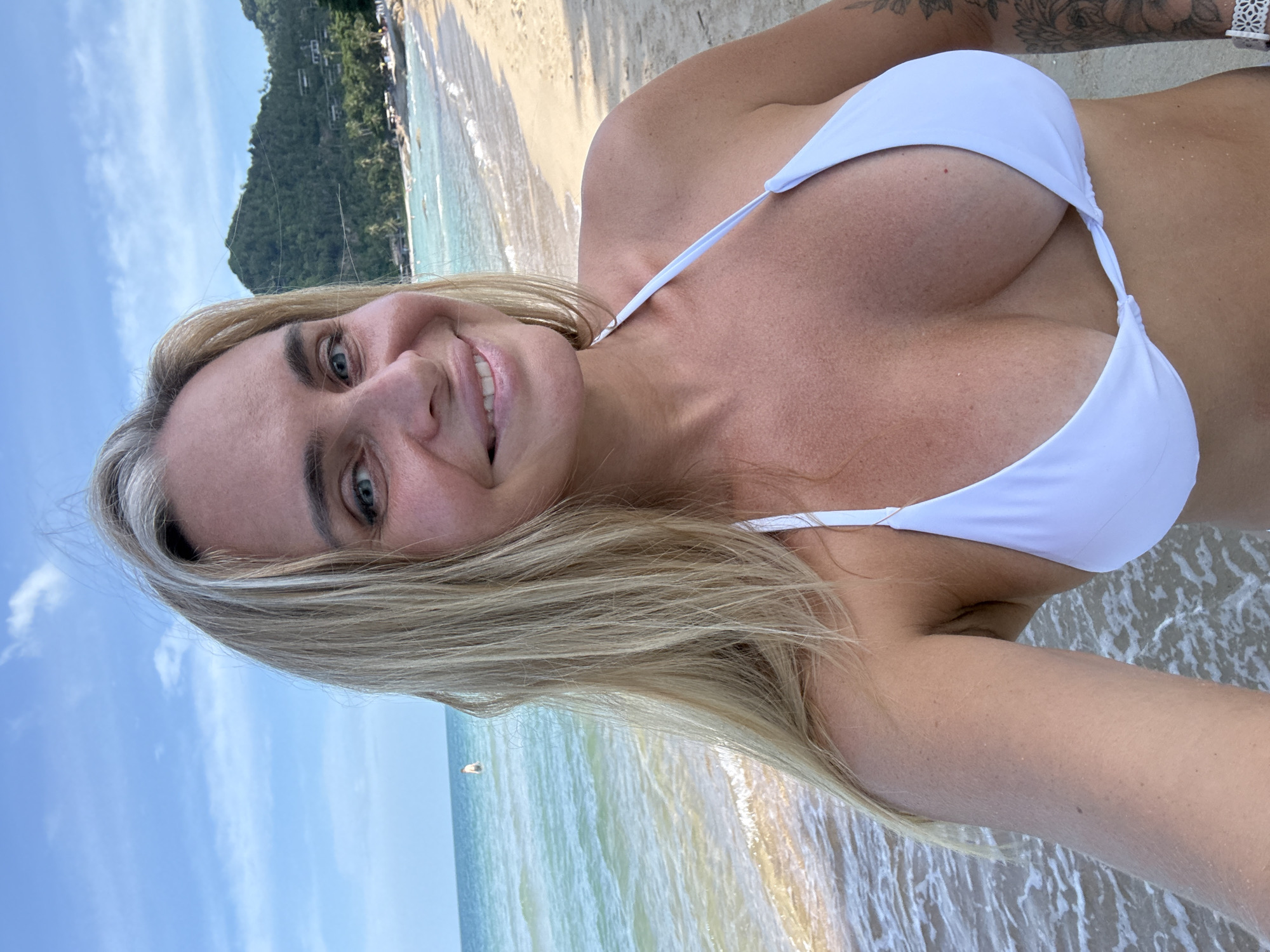 A woman with blonde hair and blue eyes smiles at the camera while standing on a beach in Ko Samui District, Thailand. She is wearing a white bikini top. The ocean is to her left, and the sand and a lush green hillside with buildings are to her right. The sky is blue with scattered clouds.