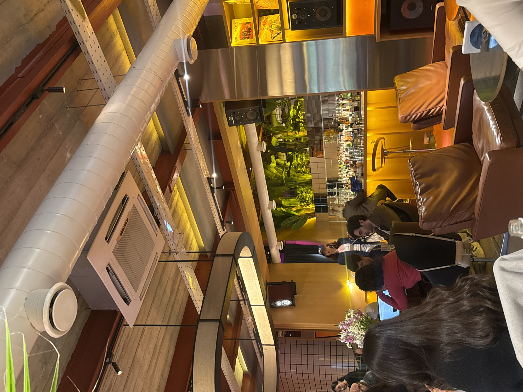 This image captures the interior of a modern and stylish cafe or lounge, featuring an industrial-chic aesthetic combined with warm, inviting elements. The ceiling showcases exposed infrastructure, including a large white circular air duct, white metal support racks, and reddish-brown beams, alongside a prominent rectangular air conditioning unit. A large, circular light fixture with a textured shade hangs from the ceiling, illuminating the space.
Several people are visible, mostly seated in comfortable brown leather armchairs and at tables. In the foreground, the back of a person with long dark hair is seen. Further into the room, a person wearing a pink top is focused on a laptop, while others are engaged in various activities, suggesting a casual co-working or social gathering.
The background reveals a well-stocked bar with numerous glass bottles, implying a bar or pub setting. Lush green indoor plants in a planter near the bar add a touch of nature. Speakers are mounted on walls and shelves, which also display framed art or record covers; the word "FOCAL" is visible on a speaker and a record cover. A vibrant floral arrangement sits on a table.
The lighting is predominantly warm and golden, creating a cozy and relaxed ambiance, characteristic of an evening setting. The overall scene depicts a lively yet comfortable environment, likely a social or study spot in Yerevan, Armenia.