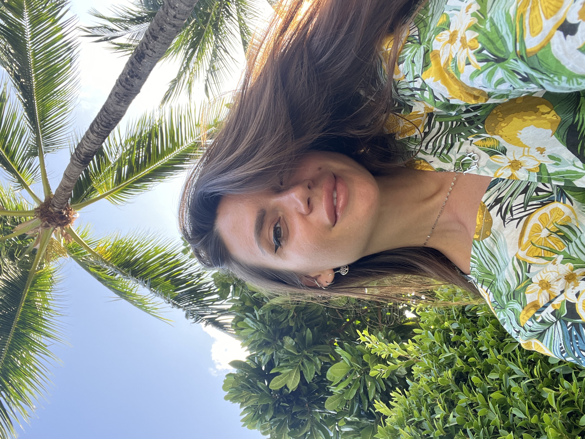 A young woman is photographed from a low angle, looking up at the camera. She has long, dark brown hair that is slightly windswept, framing her face. Her eyes are dark, with noticeable eyeliner. She is wearing a light-colored shirt with a tropical print featuring green leaves, white flowers, and yellow lemons. A delicate silver necklace with a letter "D" pendant is visible. She has small silver earrings in her left ear. Behind her, lush green foliage is visible, and the fronds of a palm tree extend over her head, with a clear blue sky visible through the leaves. The lighting suggests it is daytime, possibly late afternoon, with the sun creating a slight lens flare. The overall scene evokes a tropical or vacation setting. The location is not specified in the image.