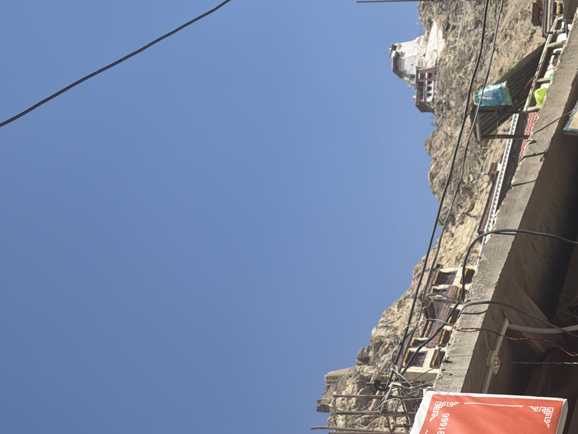The image depicts a view from a lower elevation looking up towards a settlement on a rocky hillside under a clear blue sky. In the foreground, a concrete structure with visible electrical wires and a red sign with white text are present. The red sign contains text that appears to be in a non-Latin script, along with numbers, possibly a phone number like "91666". Further up the hill, a series of buildings with white walls and red-trimmed windows are built into the rock face. At the very top of the hill, a large white structure, possibly a monastery or palace, is visible. This scene is characteristic of the city of Leh, India, known for its Tibetan Buddhist monasteries and unique architecture nestled amongst the Himalayan landscape. No people are clearly visible in the image. The overall environment suggests a sunny day in a mountainous region.