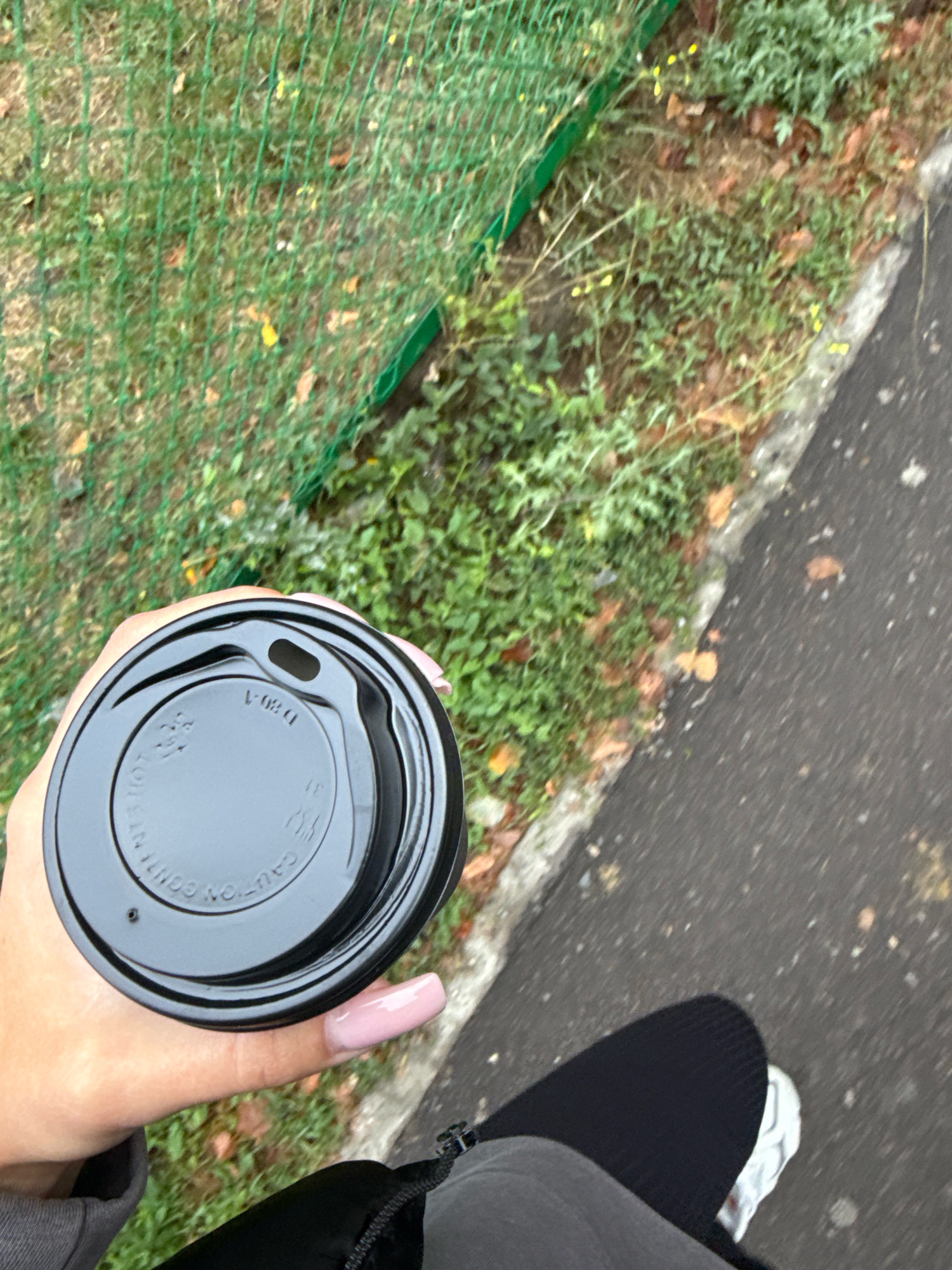 A person holds a black disposable coffee cup with a black lid while walking on a paved path next to a grassy area. The person's hand, with light pink polished nails, is visible grasping the cup. They are wearing a dark grey hooded jacket and black pants. A white sneaker is visible on their left foot. The background consists of green grass, some fallen leaves, and a green mesh fence. The scene appears to be an outdoor urban or suburban environment.