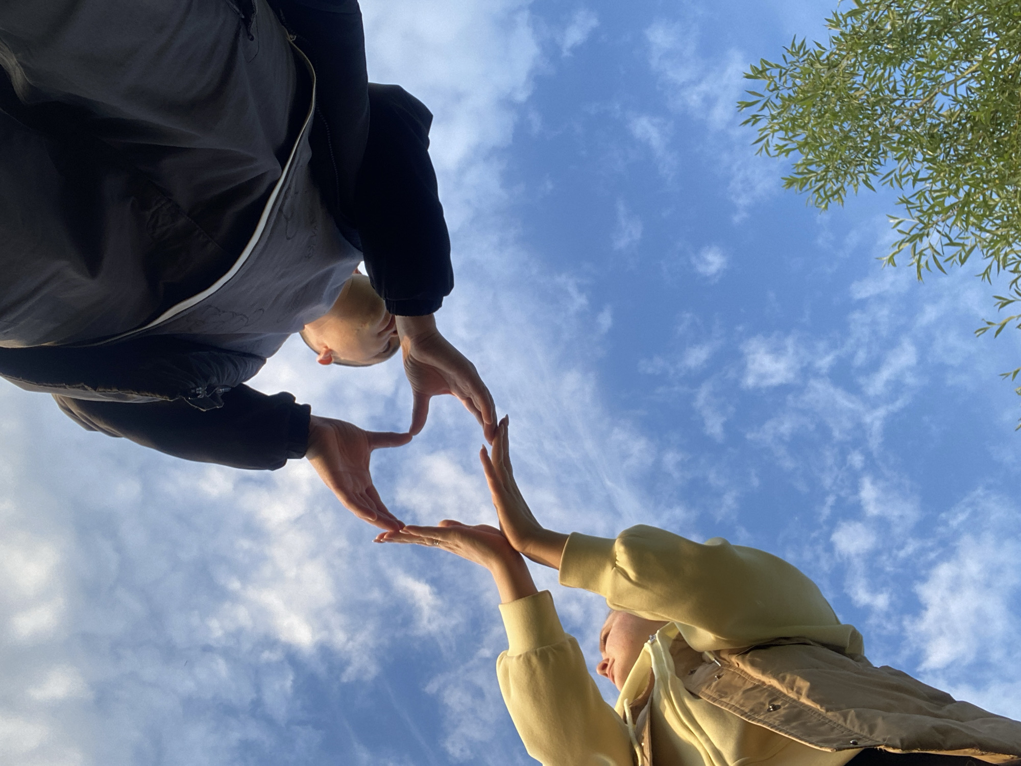 Two people are forming a heart shape with their hands against a backdrop of a blue sky with scattered clouds and a tree branch in the upper right corner. The person on the left wears a dark jacket, while the person on the right wears a yellow sweatshirt. The low-angle perspective emphasizes the sky and the hands forming the heart. The location is Chaikovsky, Russia.