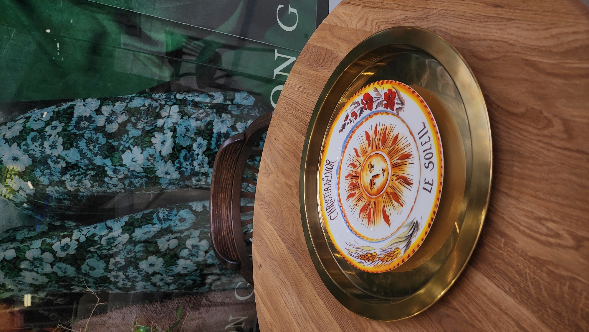 A partial overhead view reveals a round wooden table with a brass-colored metal tray resting on its surface. Inside the tray sits a decorative Christian Dior plate with "LE SOLEIL" inscribed on it, displaying a sun motif. Adjacent to the table is a window displaying floral-patterned clothing. Reflections in the glass suggest an urban setting, possibly Moscow, Russia. The image contains a detail of a chair.