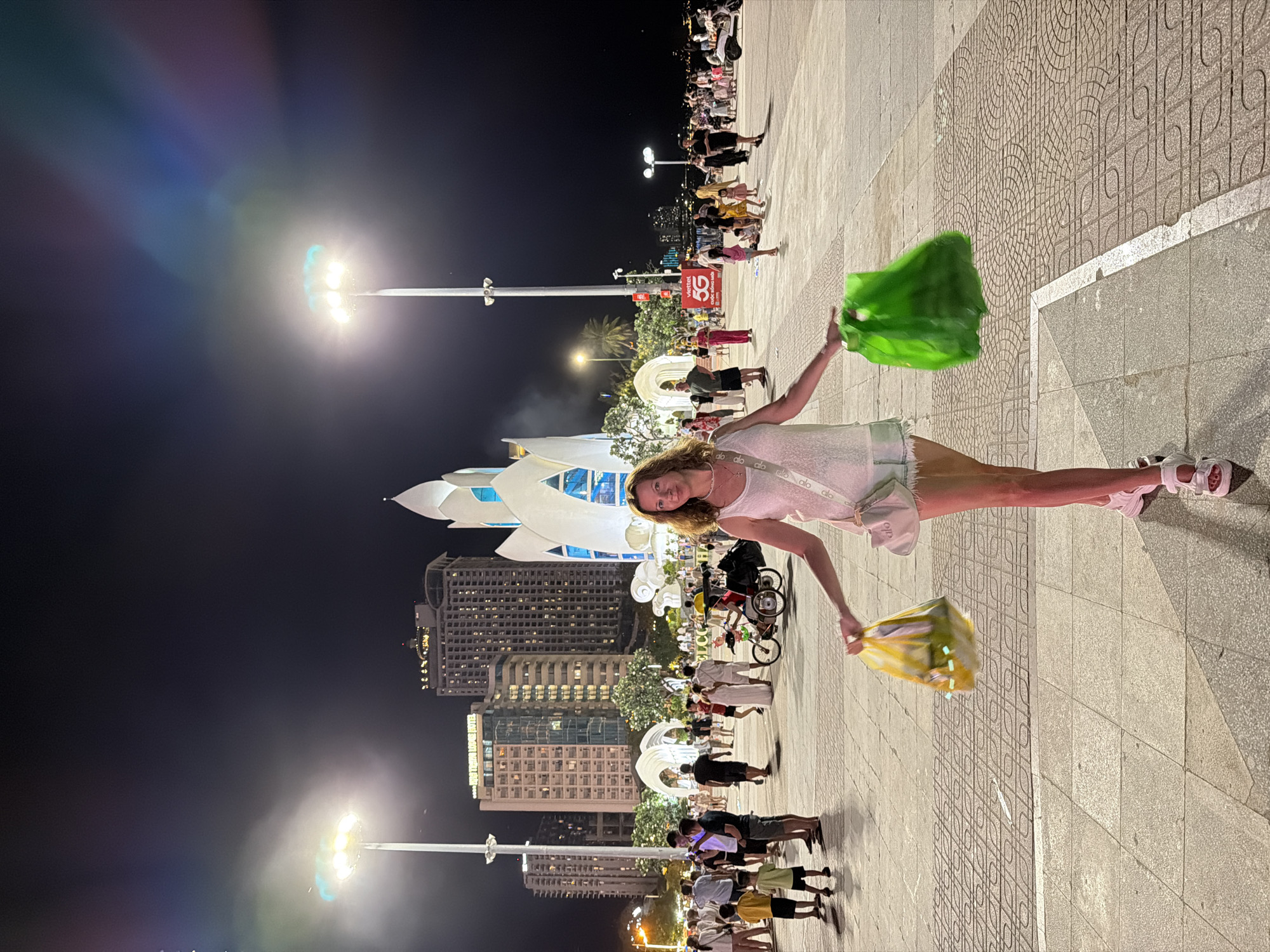 A woman is standing in the foreground, holding a yellow and a green shopping bag. She is wearing a white tank top, light-colored shorts, and white sandals. She has a white purse slung across her body. The woman is looking at the camera with a slight smile.
Behind her, the scene depicts a public square at night, likely in Nha Trang, Vietnam. Tall buildings are visible in the background, some illuminated with lights. A distinctive white, lotus-shaped building with blue accents stands out, possibly a cultural center or landmark. Streetlights illuminate the area, casting long shadows. A crowd of people are milling about, some walking, others standing in groups. A bicycle with a sidecar is also present, with two people in it. A sign with "5G" is visible on a building in the mid-ground. The ground is paved with interlocking tiles. The atmosphere appears festive or busy, typical of a public gathering space in a city at night.