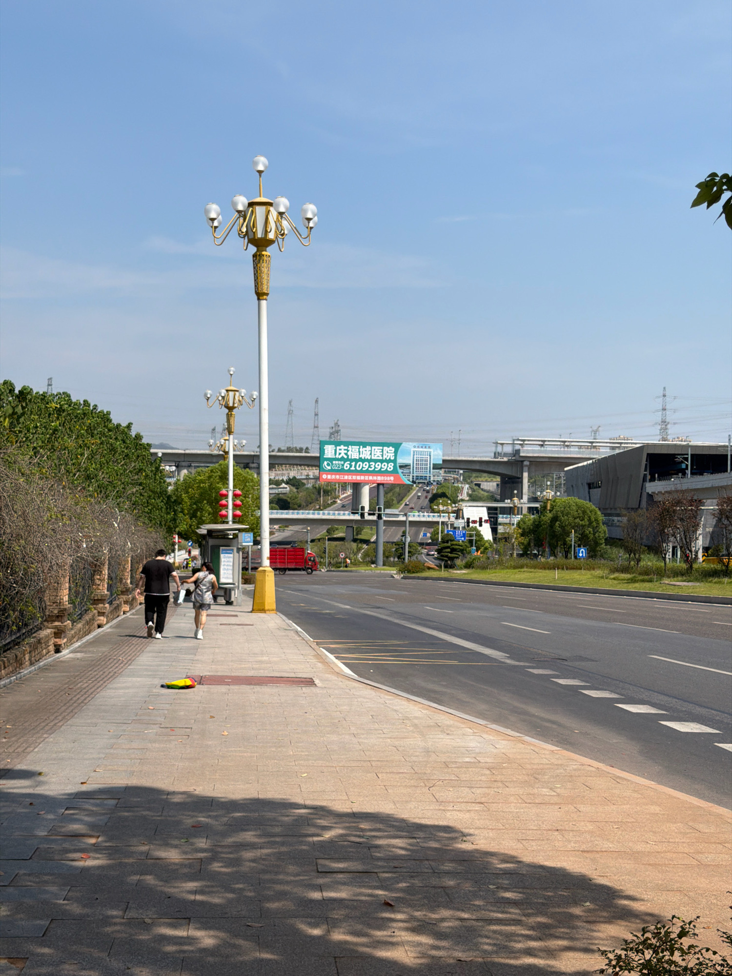 The image depicts a street scene in the Jiangjin District of Chongqing, China, on a sunny day. In the foreground, a pedestrian walkway made of light brown interlocking tiles runs alongside a multilane asphalt road. Two individuals, a man in a black t-shirt and black pants and a woman in a light-colored tank top and shorts, are walking away from the camera on the walkway. To the left of the walkway, a brick wall with a metal fence runs along a strip of trees and bushes. A tall, ornate street lamp with multiple frosted globes stands prominently on the walkway. Further down the walkway, a bus stop shelter with a blue roof is visible, and a red vehicle, possibly a truck, is seen on the road in the distance. A large billboard with Chinese text and an image of a building is mounted on a pole in the middle ground, partially obscured by an elevated road structure. The sky is clear and blue.