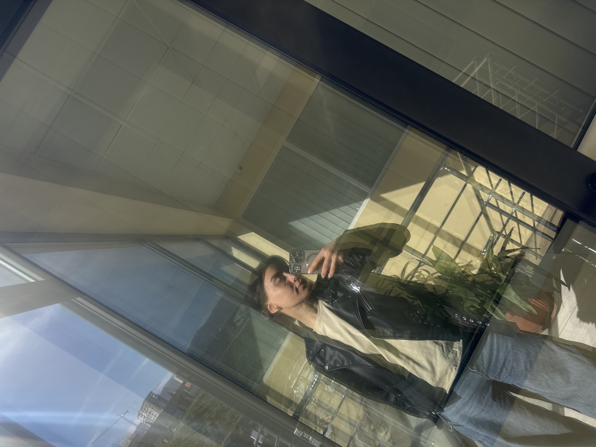 A person is taking a selfie in front of a large window. The reflection shows a view of a building under construction and the sky. The person is wearing a black jacket, a light-colored shirt, and jeans. Reflected behind the person is the interior of a building, including a potted plant and a balcony with a railing. The location is identified as Tbilisi, Georgia.