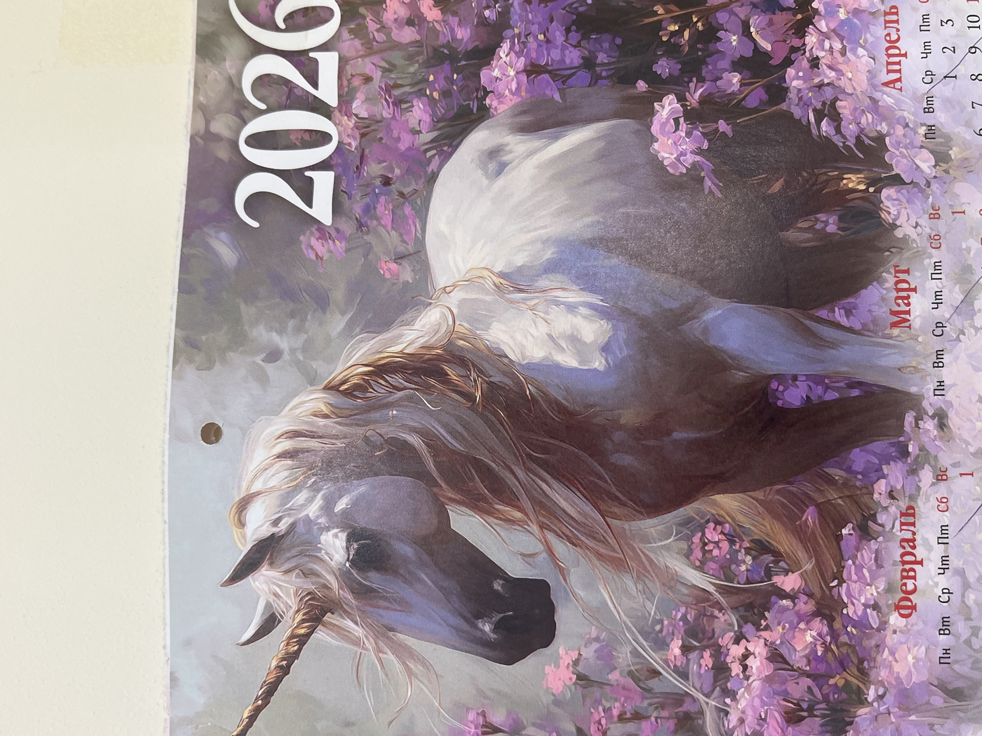 The image is a photograph of a calendar page featuring a painting of a white unicorn with a golden horn, surrounded by purple flowers. The year "2026" is prominently displayed in white text. Below the unicorn painting, the months of February, March, and April are labeled in Russian, with corresponding days of the week abbreviations and a portion of the calendar grid visible. The image does not contain any information about the city of Ufa, Russia, or any specific location. There are no people or discernible actions in the image.