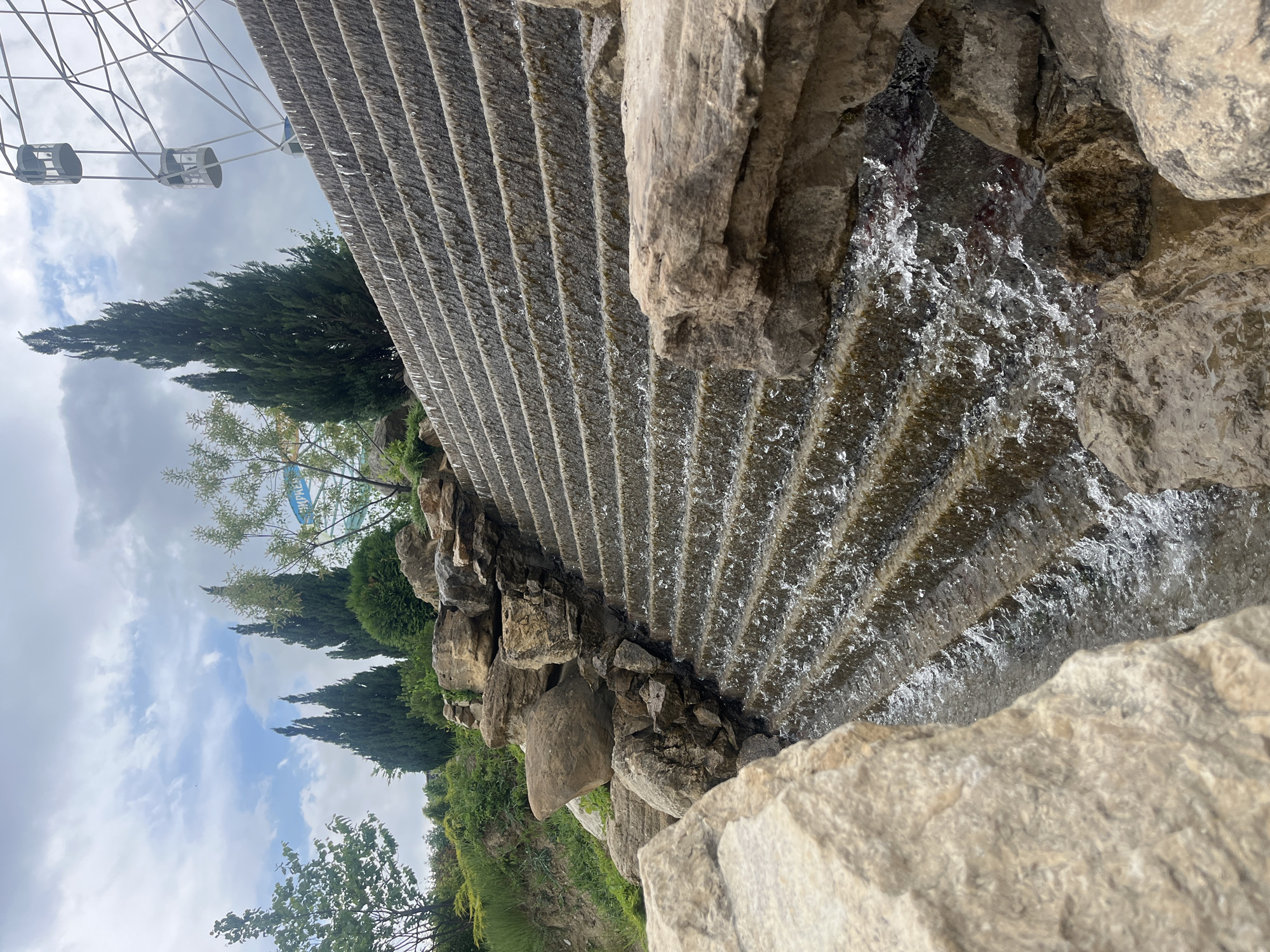 The image depicts a tiered waterfall flowing down a stone structure in Novo-Zubutli, Russia. The water cascades over multiple steps, framed by large, light-colored rocks in the foreground. Evergreen trees and other greenery are visible in the background, along with a Ferris wheel on the left. The sky is overcast with cloudy conditions.